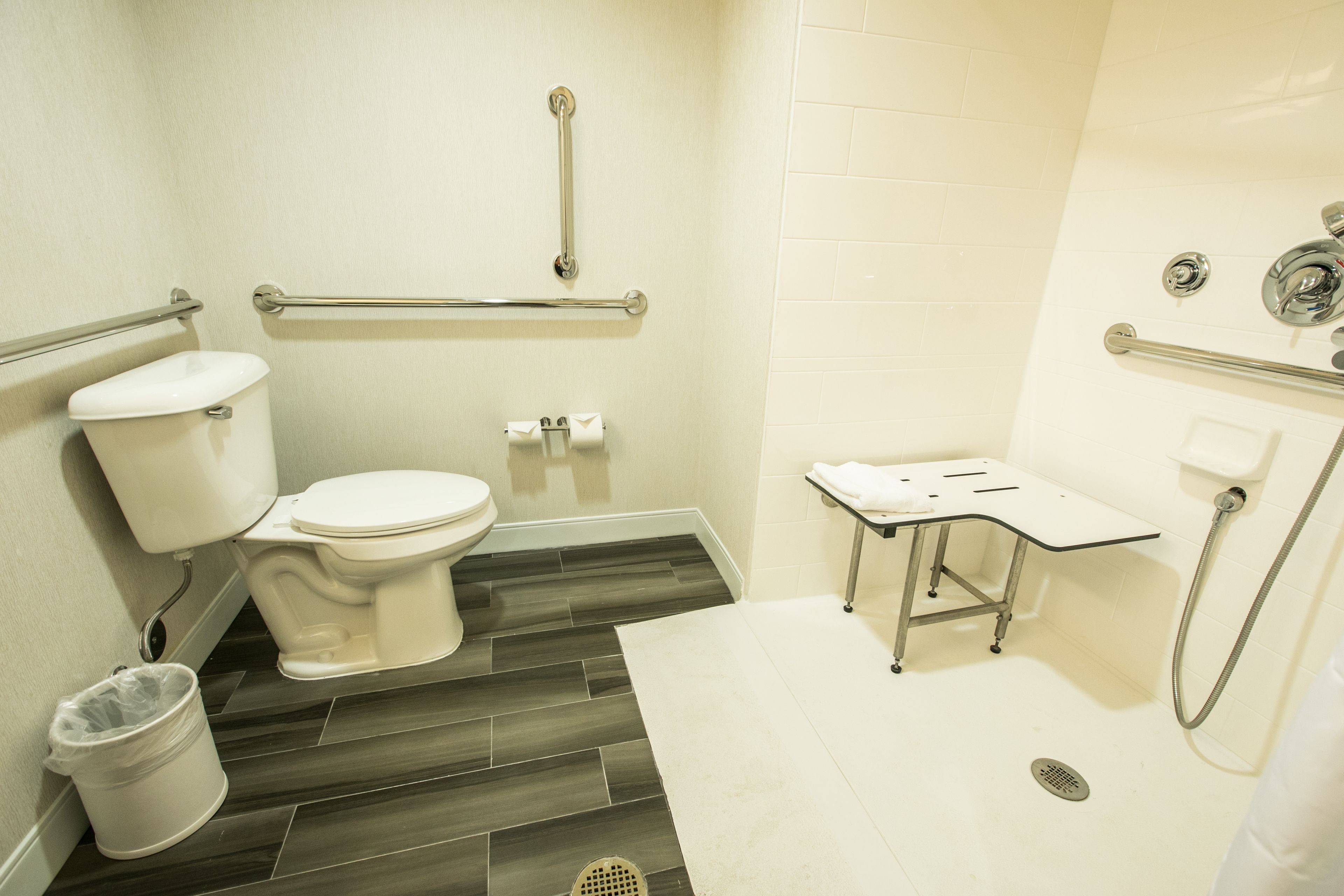 A bathroom with a toilet, grab bars, a shower area, and a trash can.
