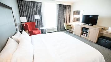 Et hotelværelse med en stor seng, et skrivebord med et fjernsyn, en rød stol og et vindue med gardiner.
