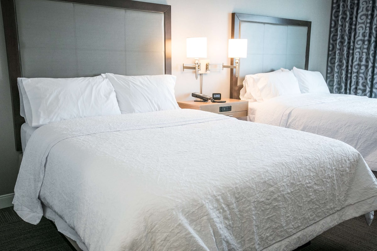 A hotel room with two beds, each with white linens and headboards. There are bedside tables with lamps and a clock.