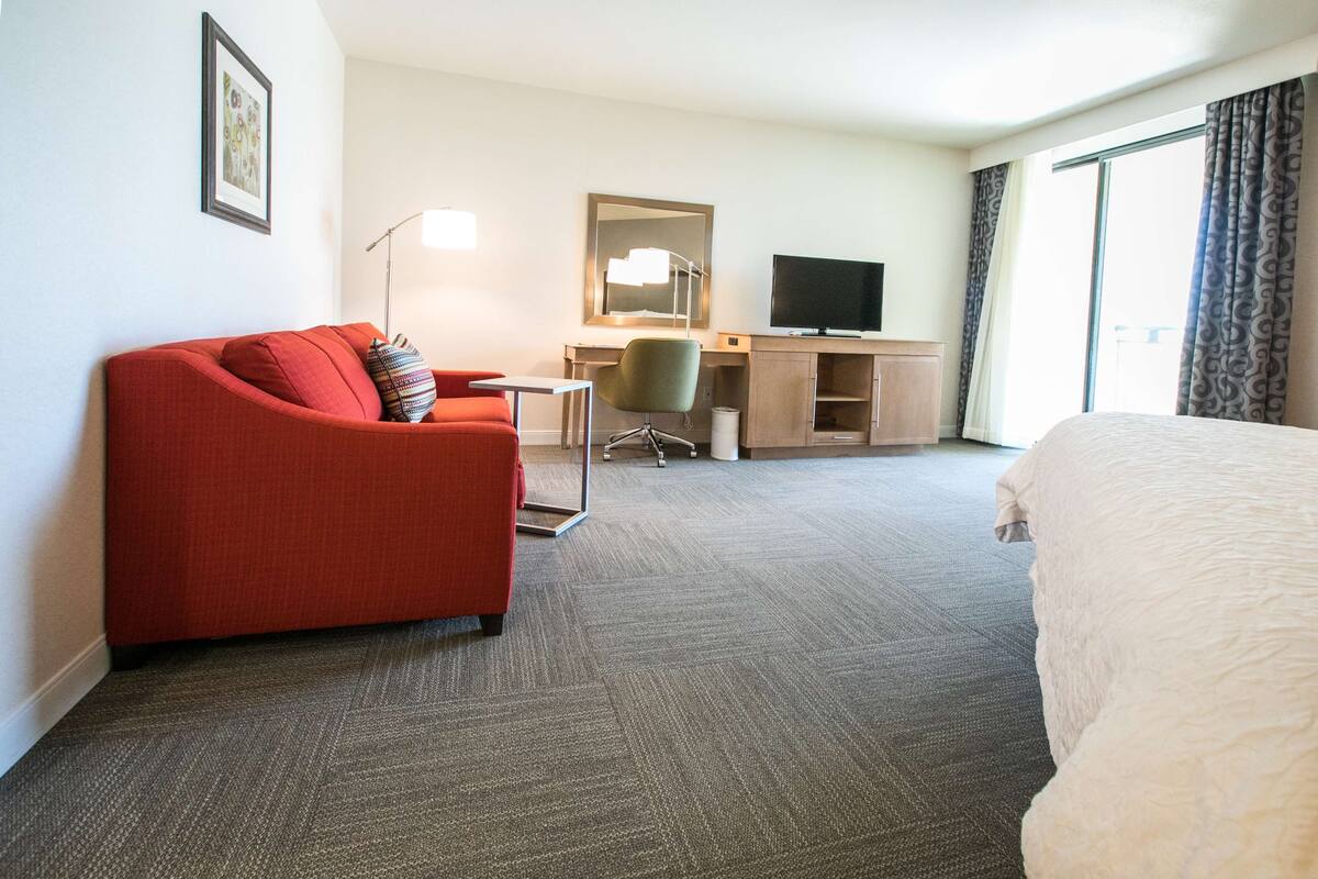 A hotel room with a red sofa, a bed, a desk with a computer, a TV, and a large window with curtains.