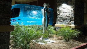 A blue bus with "It's a day for good" and "hampton.com" on it, parked in front of a building with a stone wall and palm plants.