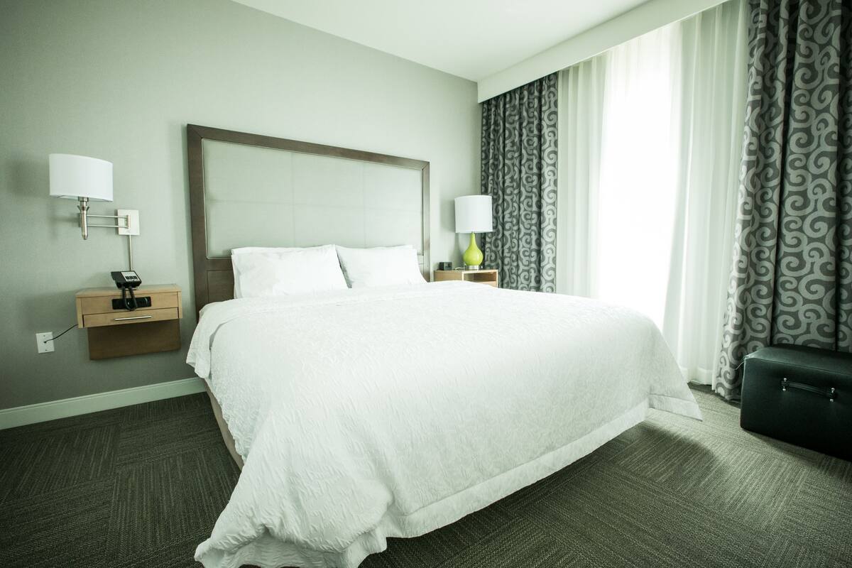 A hotel room with a large bed, two bedside tables, a lamp, a suitcase, and a patterned curtain.