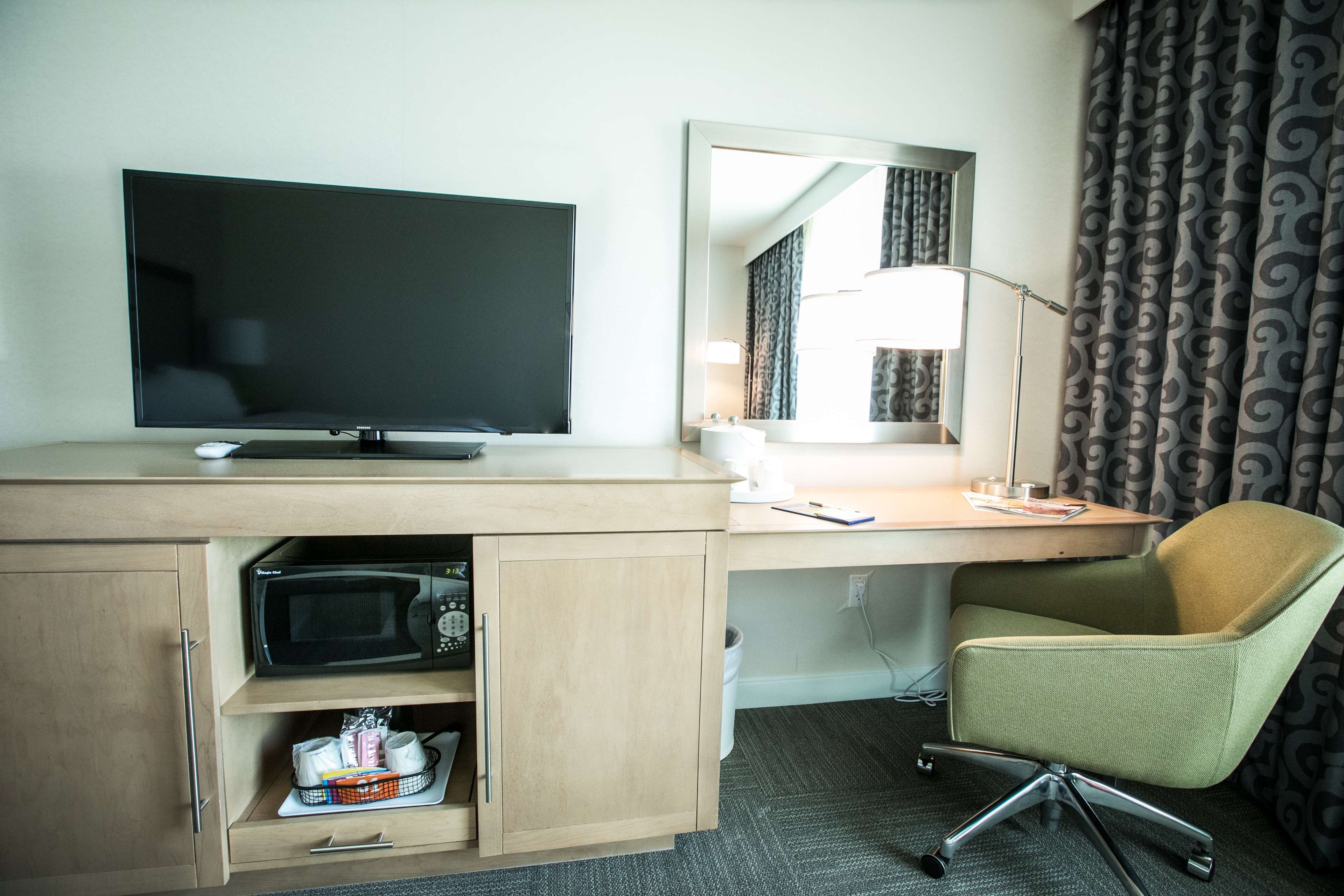 A hotel room with a flat-screen TV, a desk with a lamp, a green office chair, and a microwave in a wooden cabinet.