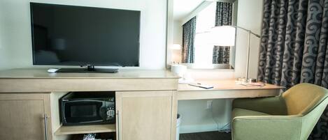 A hotel room with a flat-screen TV, a desk with a lamp, a green office chair, and a microwave in a wooden cabinet.