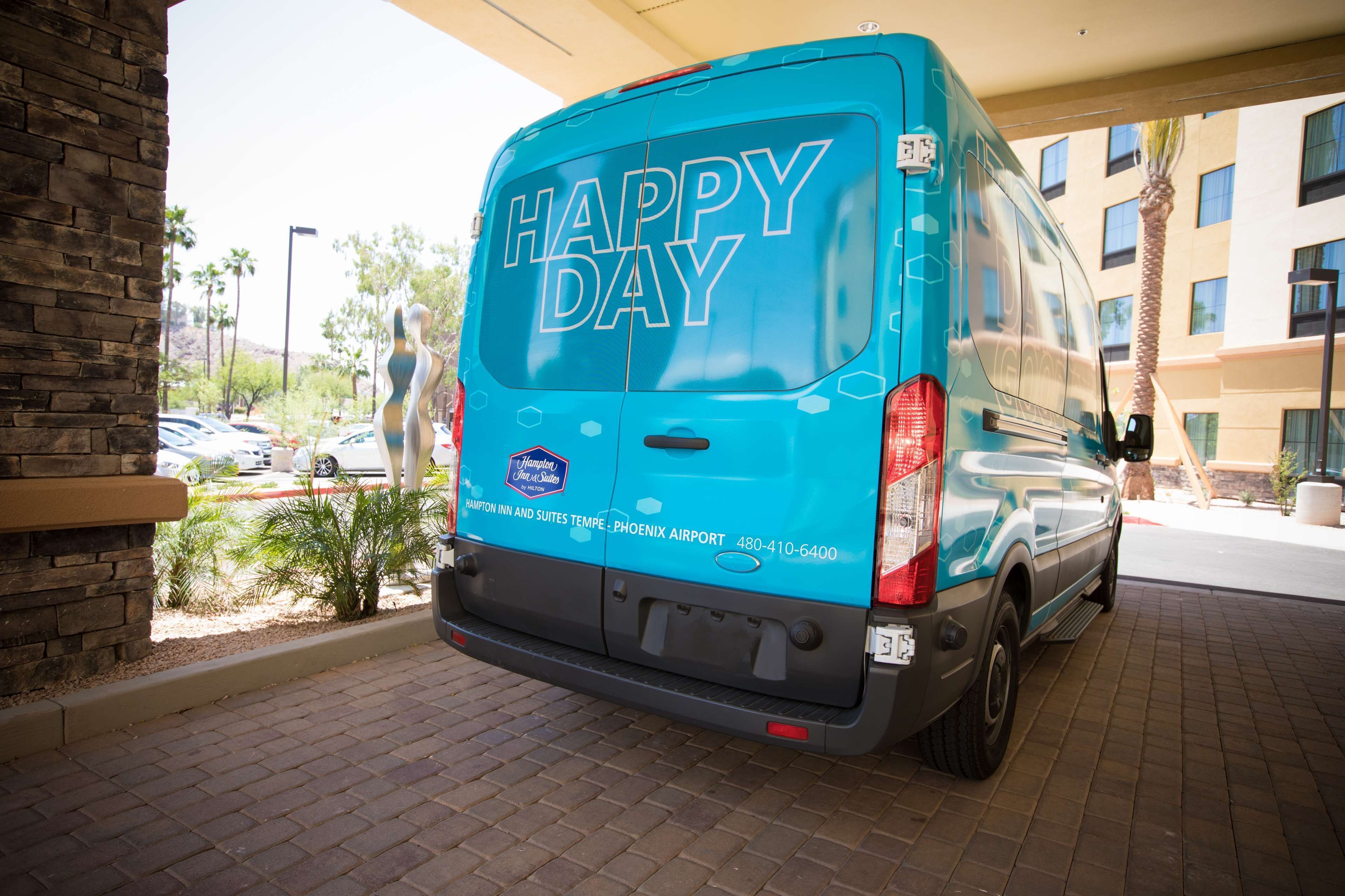 A blue van with "HAPPY DAY" and "Hampton Inn and Suites Tempe - Phoenix Airport" on the back, parked in a lot.