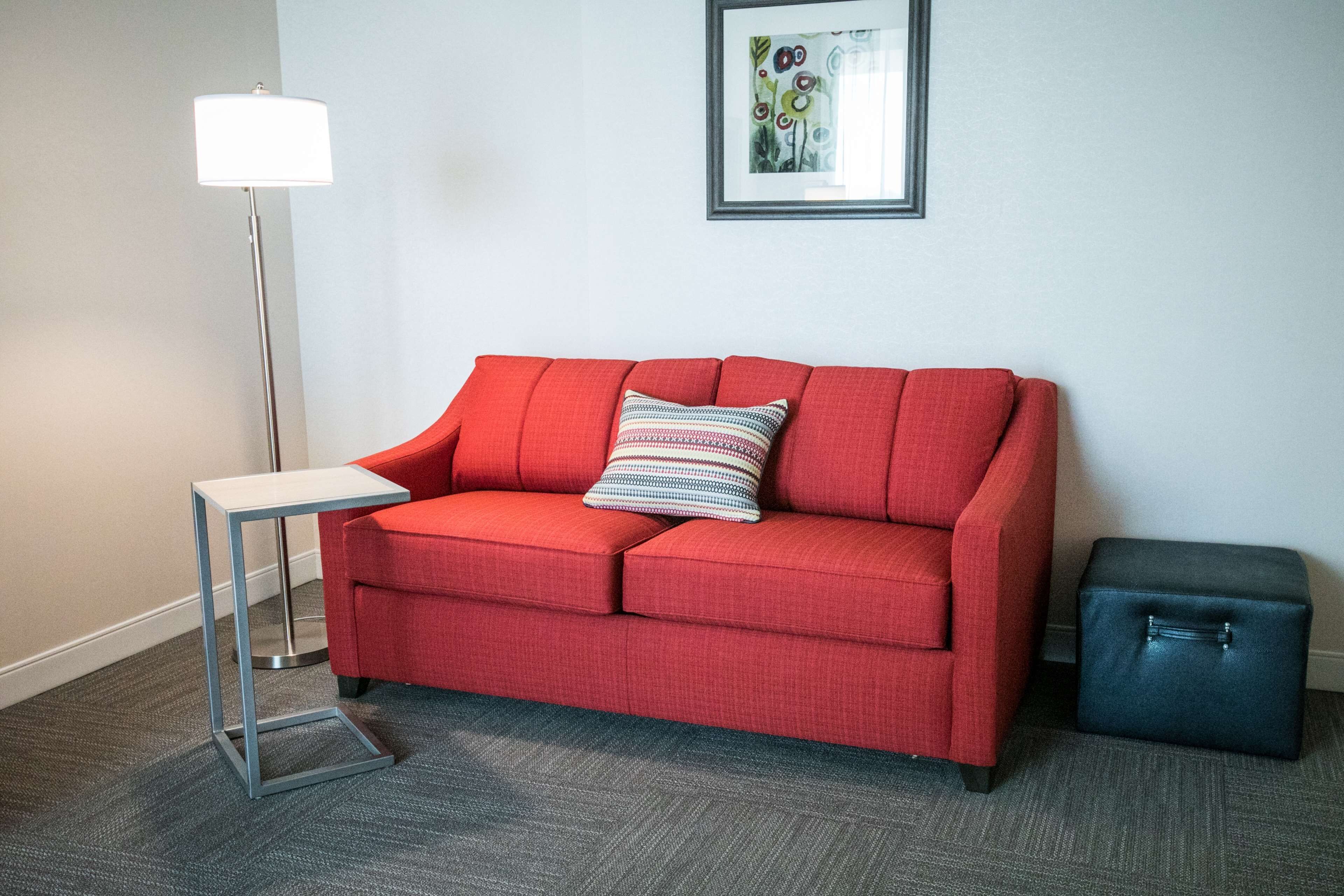 Un canapé rouge avec un coussin à rayures, une petite table d’appoint blanche, un pouf noir et une lampe sur pied.