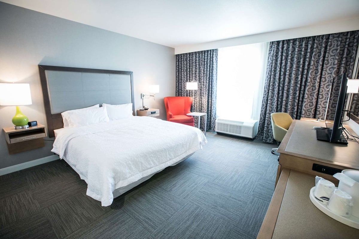 A hotel room with a large bed, a desk with a computer, a chair, a lamp, and a window with curtains.