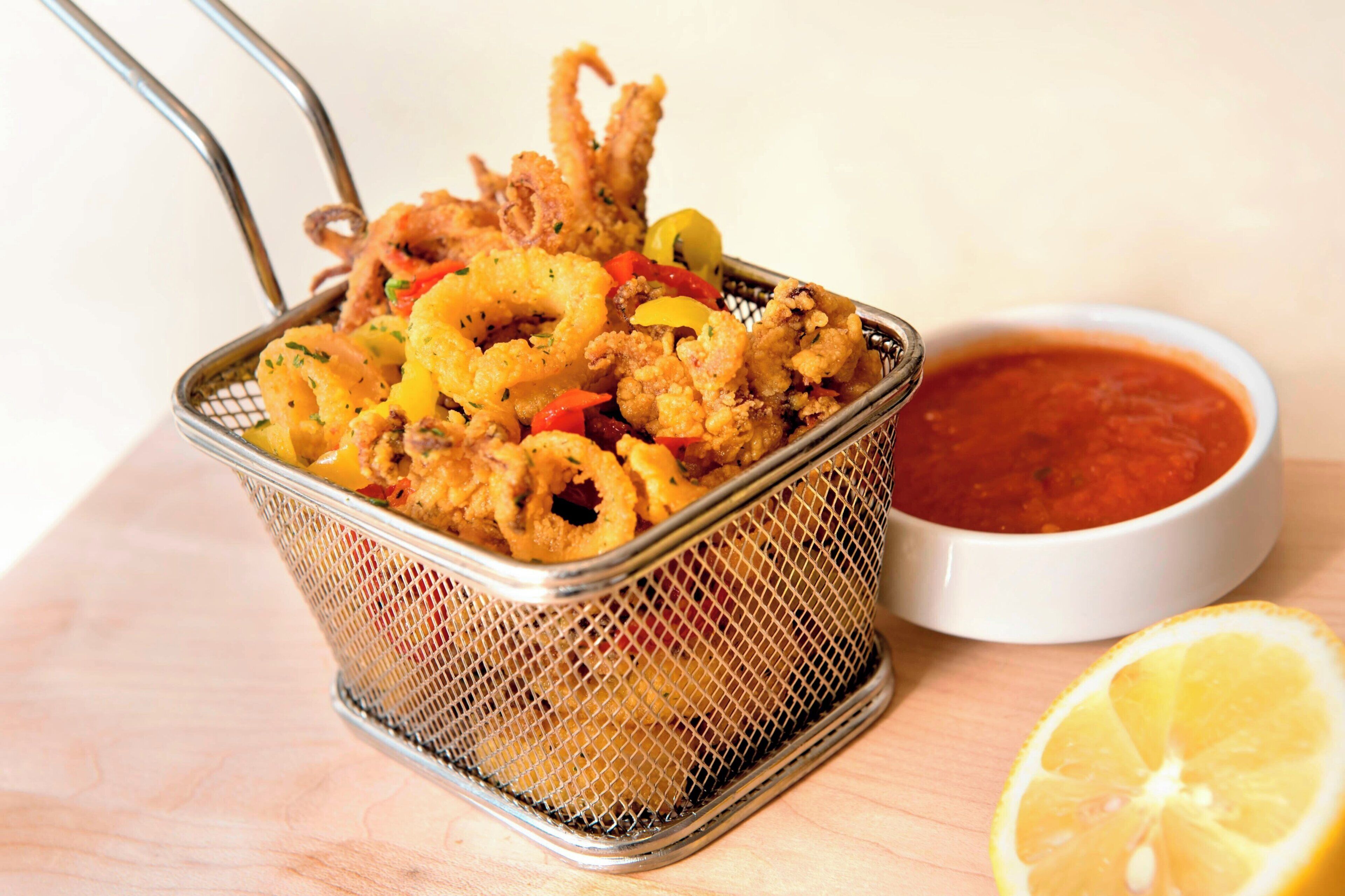 Una cesta de calamares fritos, acompañada de salsa marinara y un gajo de limón.
