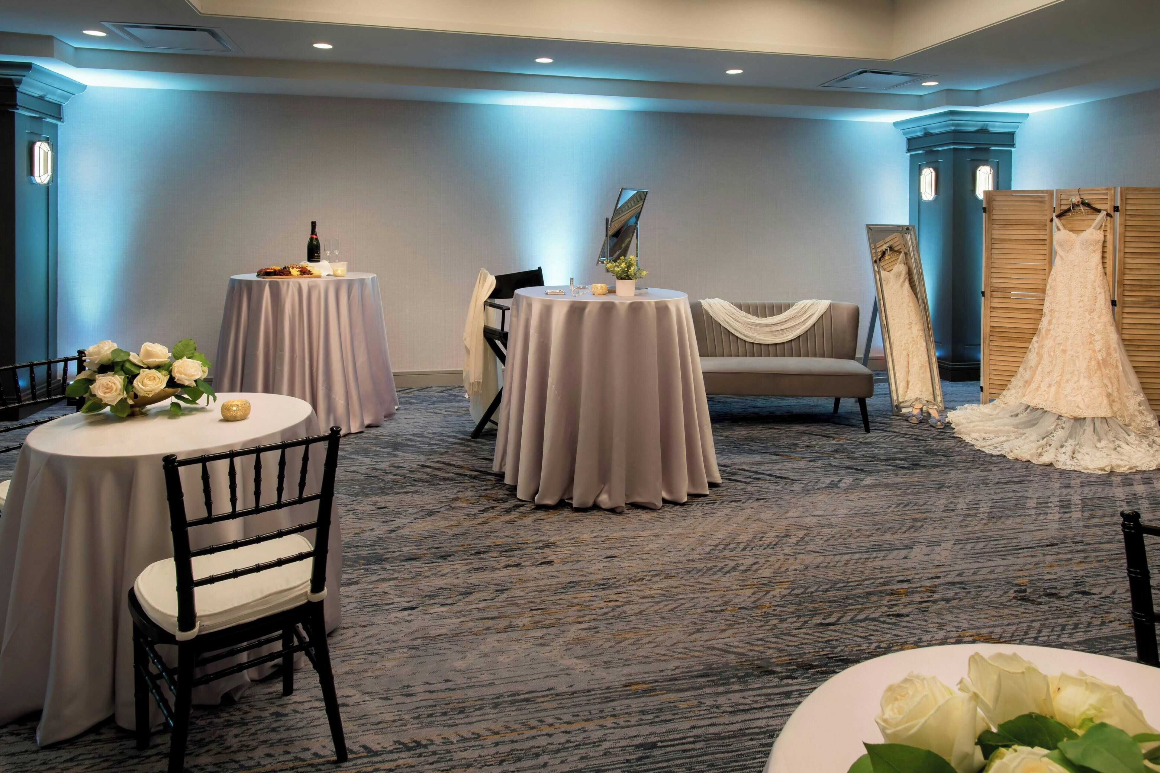 Habitación de hotel con dos mesas redondas, un sofá y un vestido de novia exhibido.