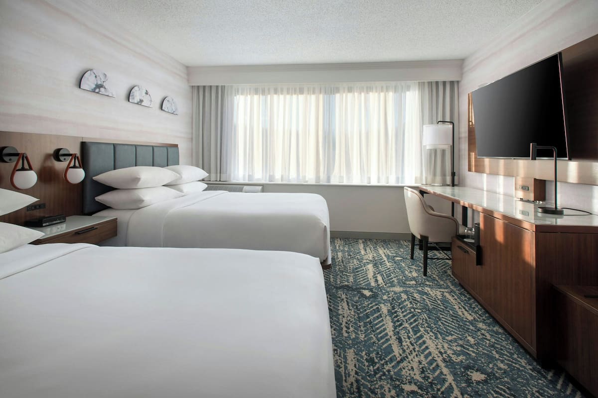 A hotel room with two beds, a desk, a TV, and a large window with curtains.