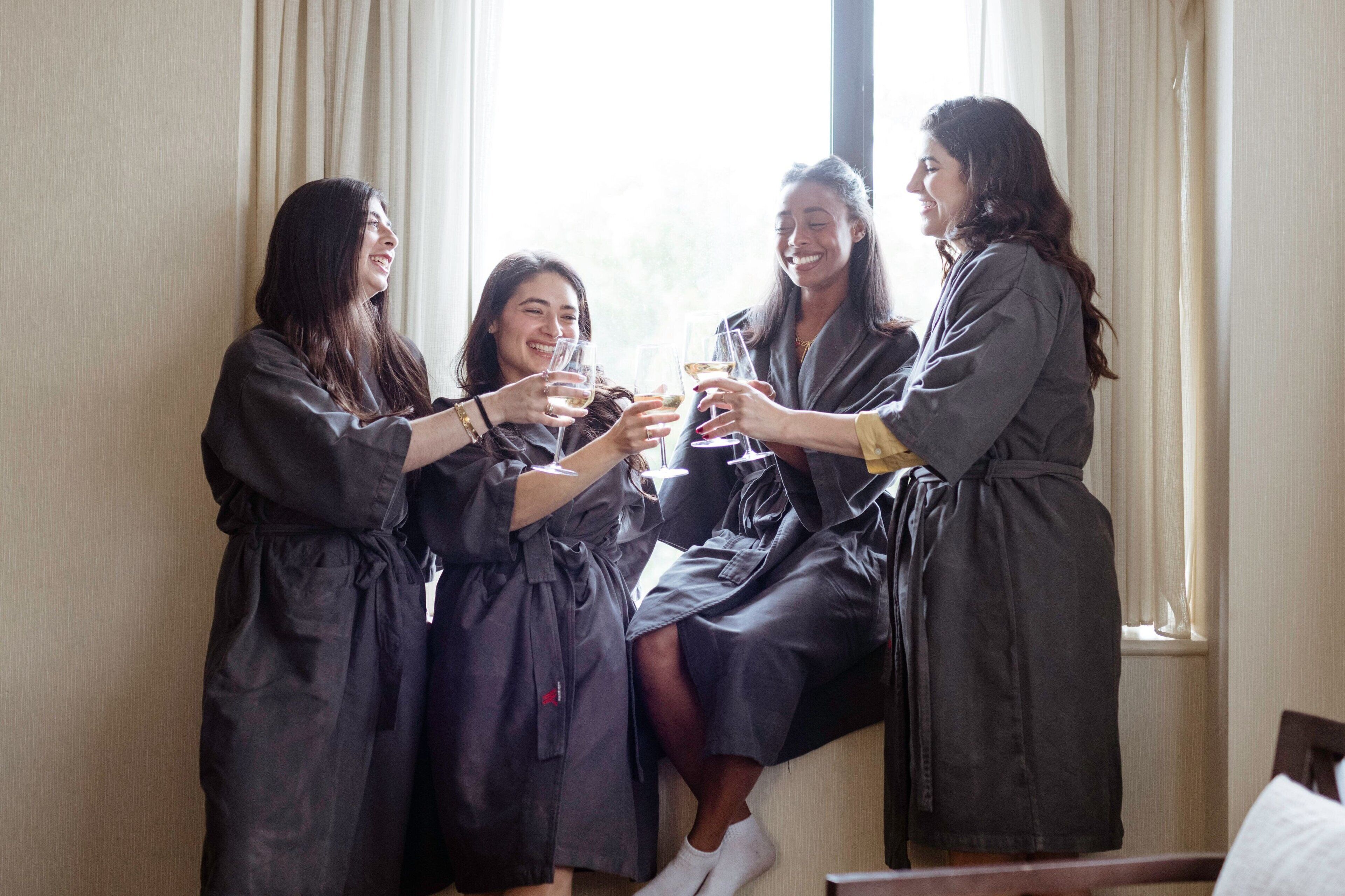 Four individuals in dark robes toasting with wine glasses near a window with curtains.