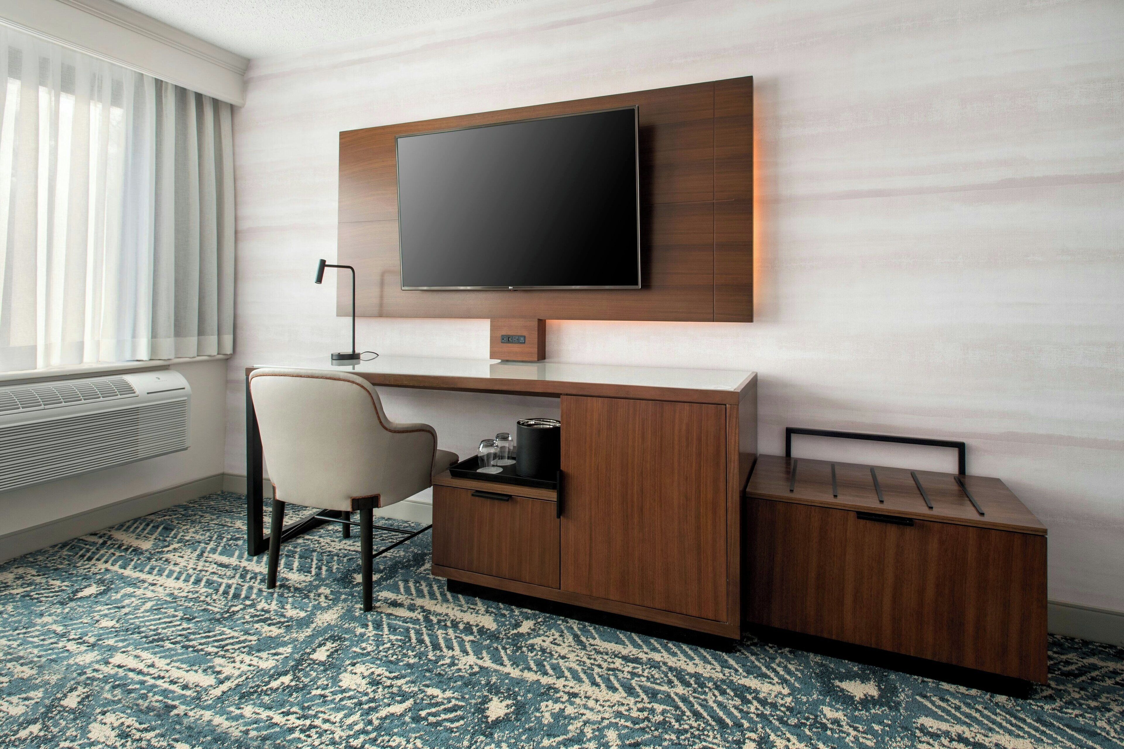 Habitación de hotel con televisión de pantalla plana montada en la pared, un escritorio de madera con una silla y una alfombra estampada.