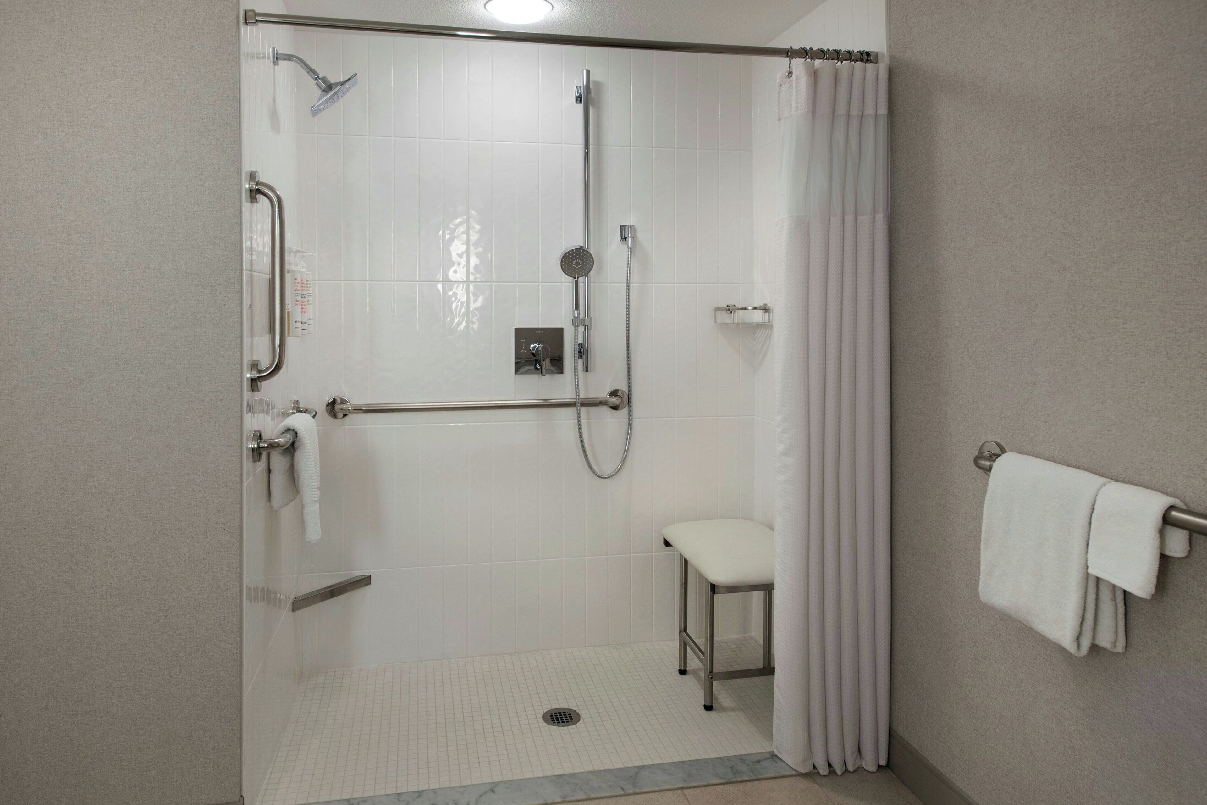 A shower with a glass door, a handrail, a showerhead, a handheld showerhead, a towel rack, and a white chair.