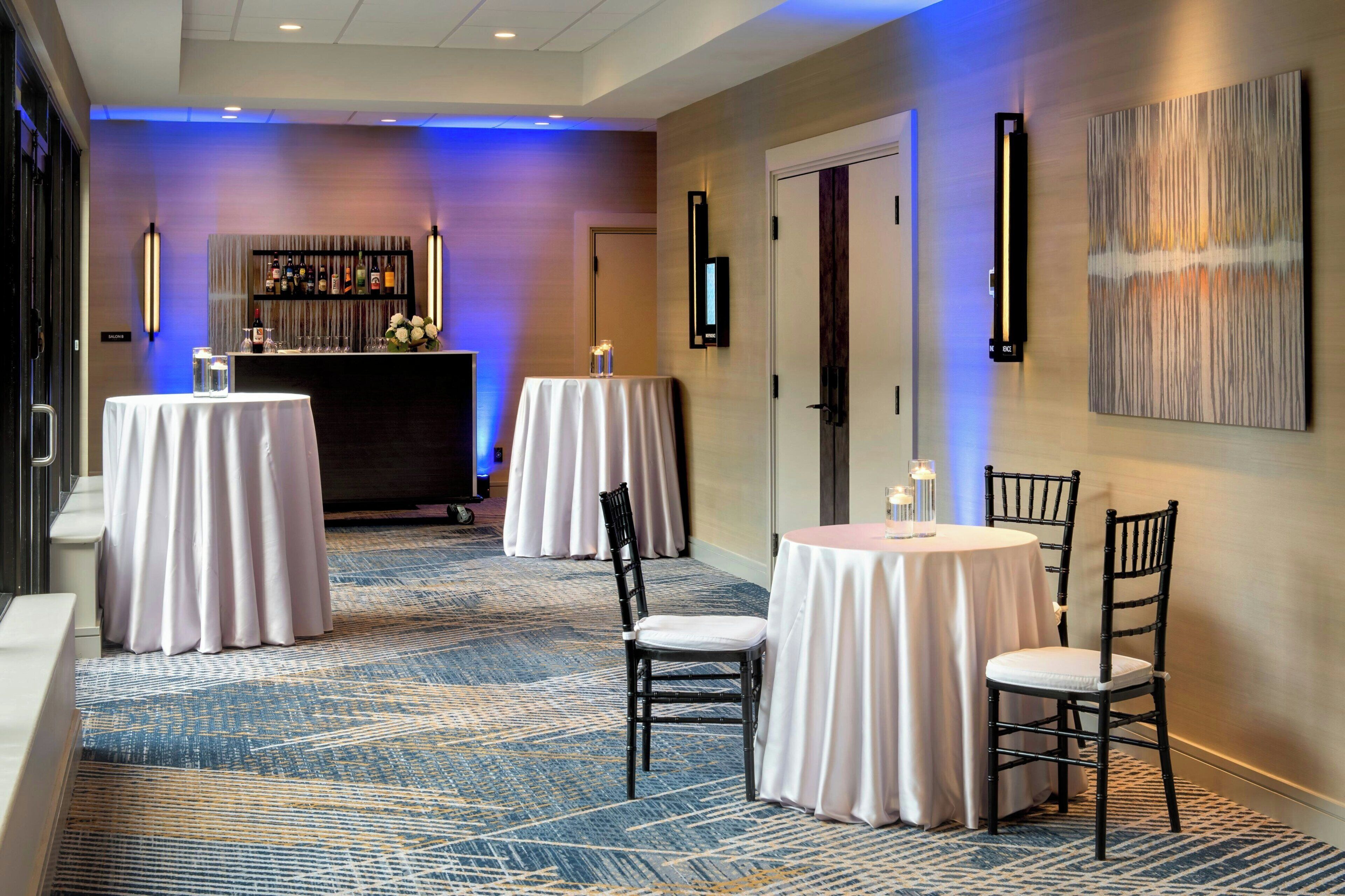 A hotel lobby with a bar area, two round tables with chairs, and a patterned carpet.