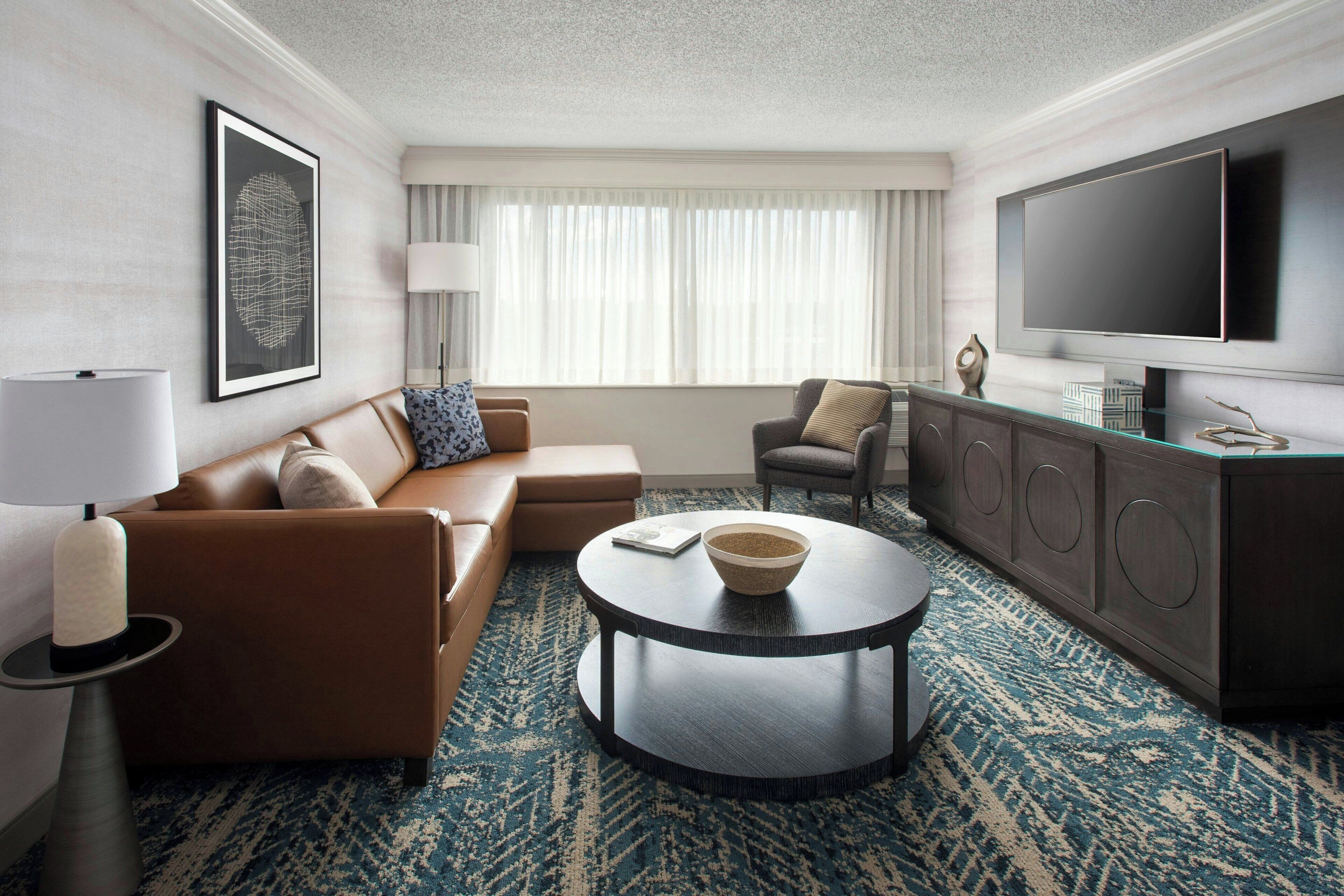 Une chambre d’hôtel moderne avec un canapé en cuir, une table basse ronde, un téléviseur et une grande fenêtre avec des rideaux.