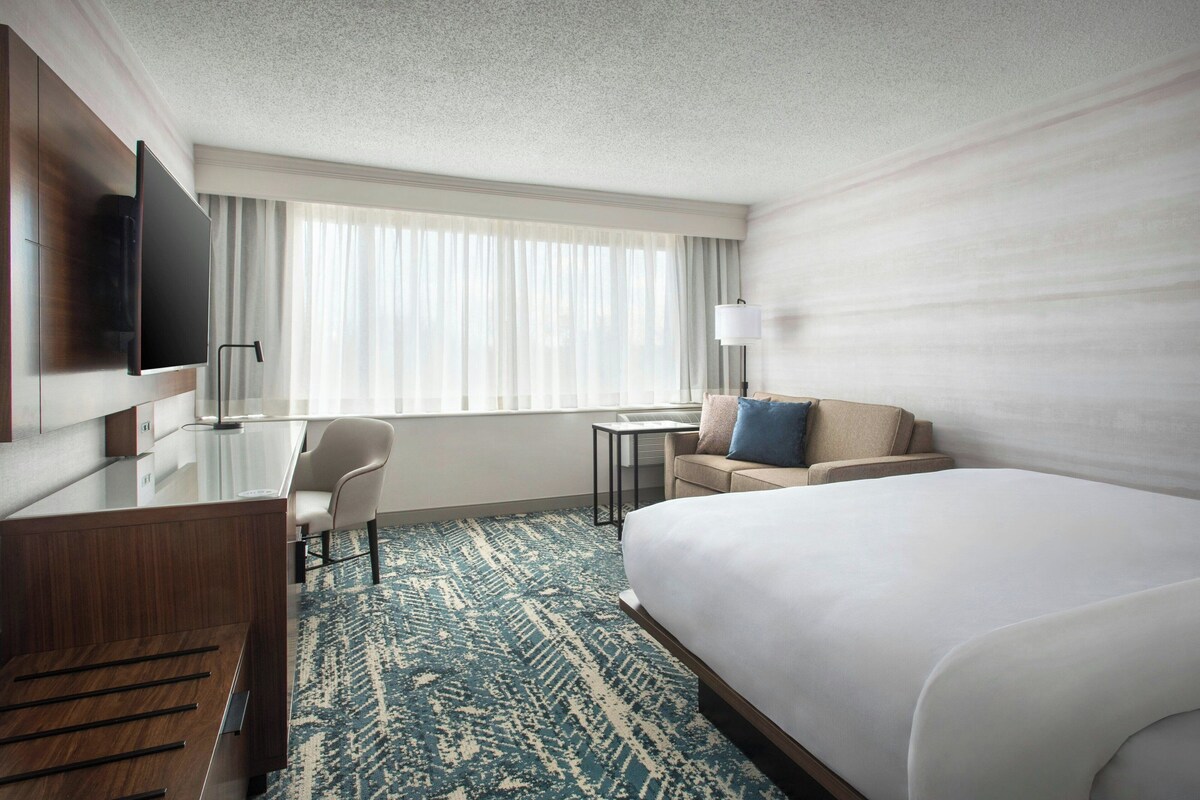 Habitación de hotel con cama, escritorio, silla, televisor y ventana con cortinas.