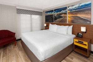 Un cuarto de hotel con una cama grande, una silla roja, una mesita de noche con una lámpara y tres cuadros en la pared.