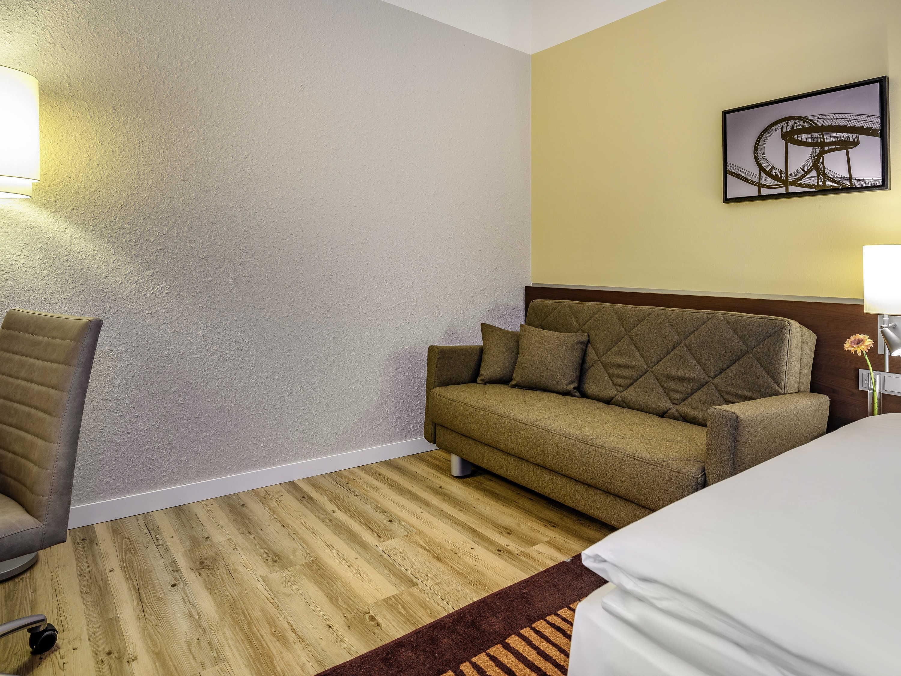 A hotel room with a wooden floor, a sofa, a chair, a bed, and a wall-mounted lamp.
