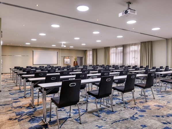 Une salle de conférence avec des rangées de tables et de chaises, un projecteur, un tableau blanc et une grande fenêtre avec des rideaux.