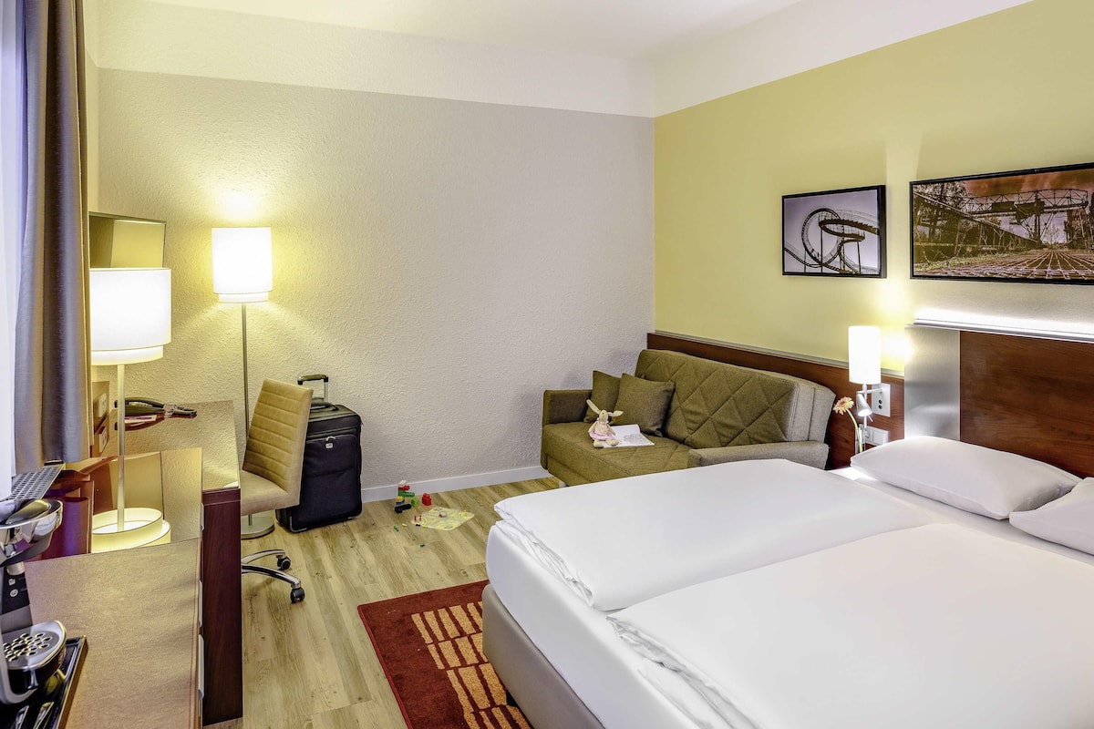 Een hotelkamer met een bed, een bureau met lamp, een stoel, een koffer en een kleine tafel met een lamp.