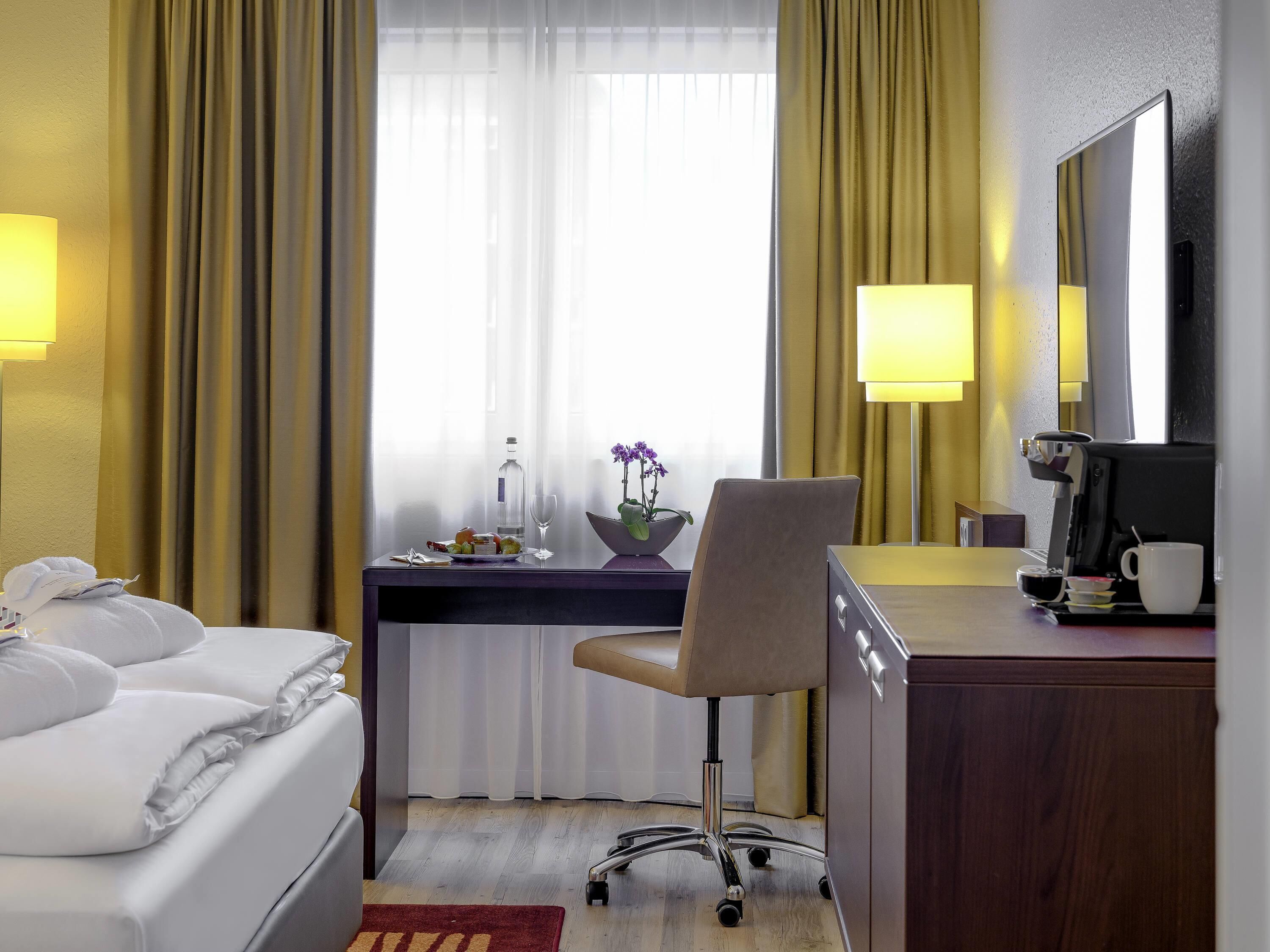 A hotel room with a bed, desk, chair, coffee machine, and a window with curtains.