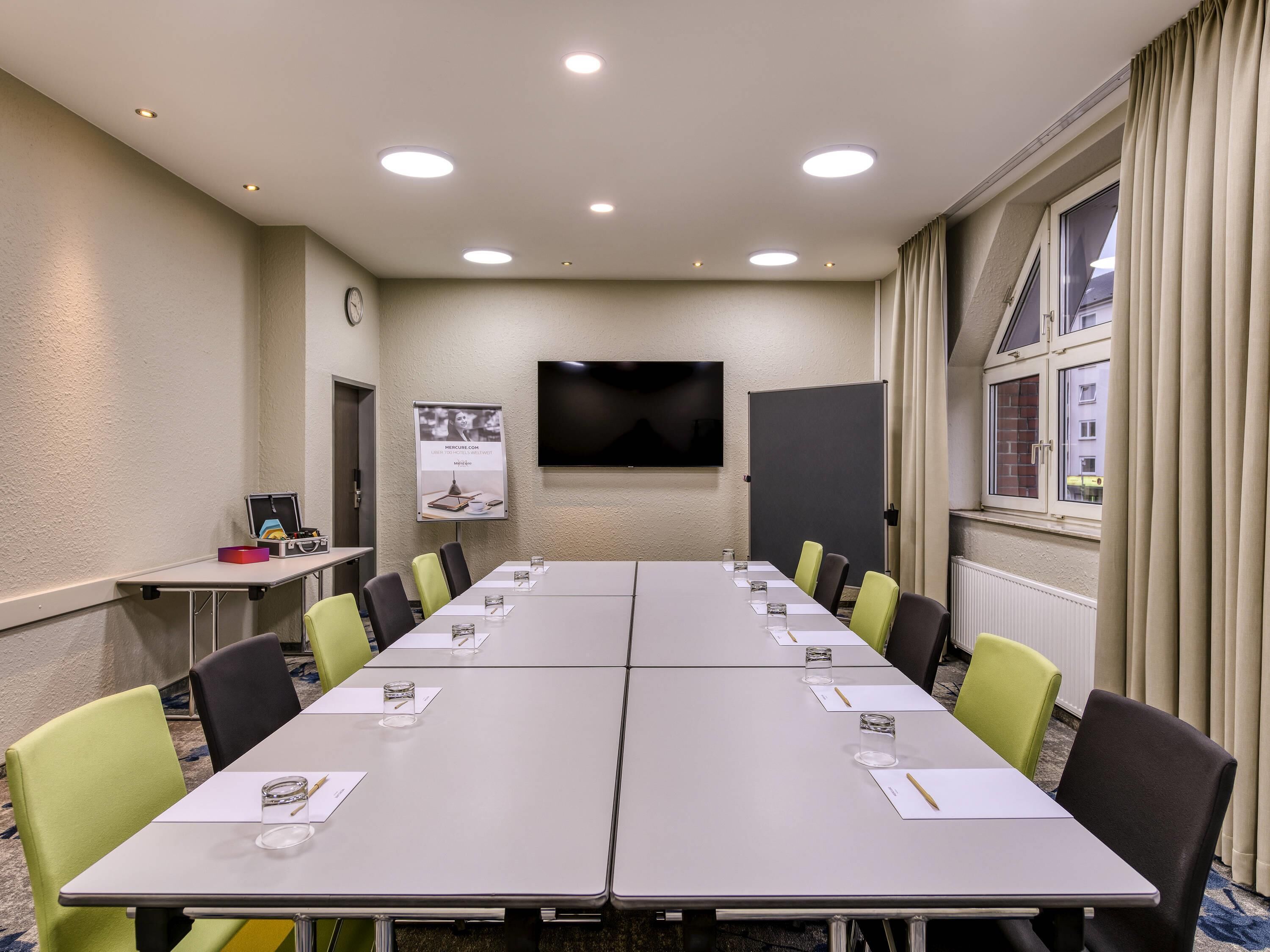Un salón de conferencias con una larga mesa, sillas, un reloj, una pantalla de proyector y una ventana con cortinas.