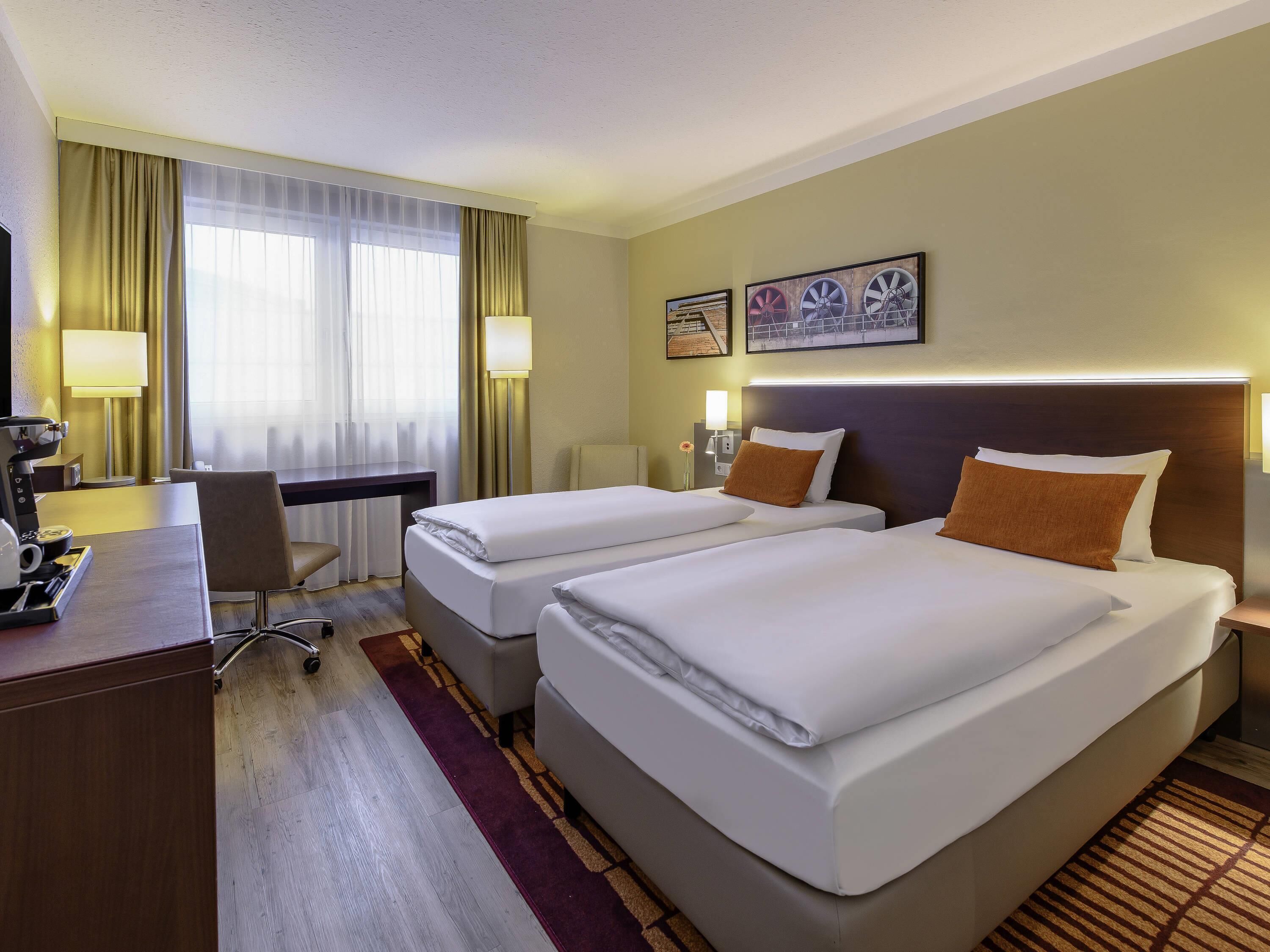 A hotel room with two beds, a desk, a chair, a lamp, and a window with curtains.
