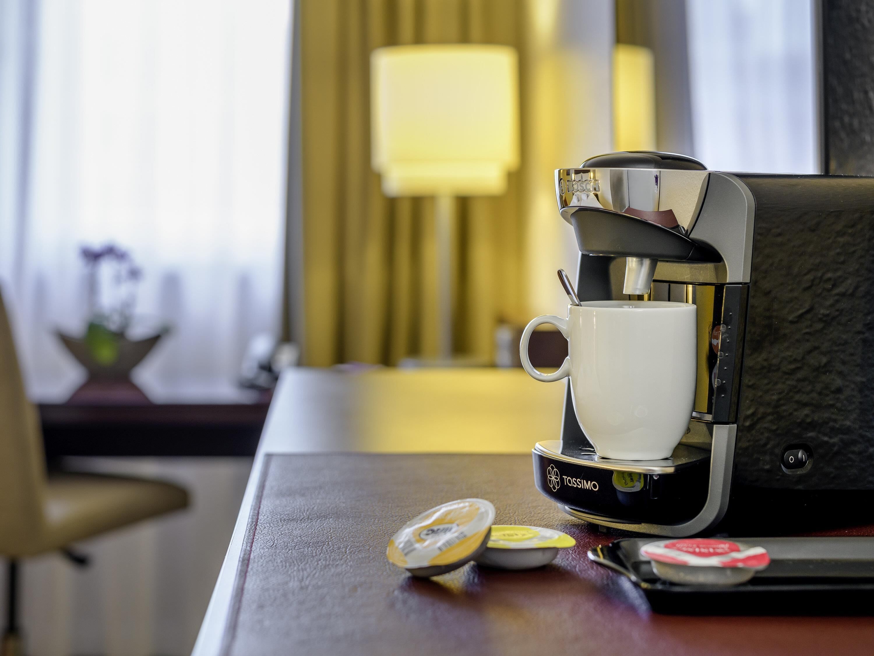 Una cafetera con una taza sobre la encimera, una lámpara al fondo y una silla.