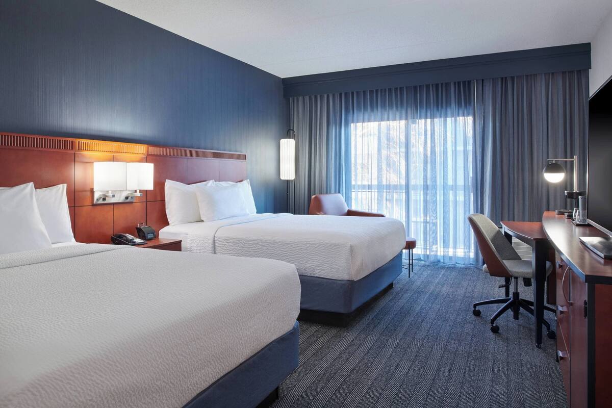 A hotel room with two beds, a desk, and a chair. There is a window with curtains and a lamp on the desk.