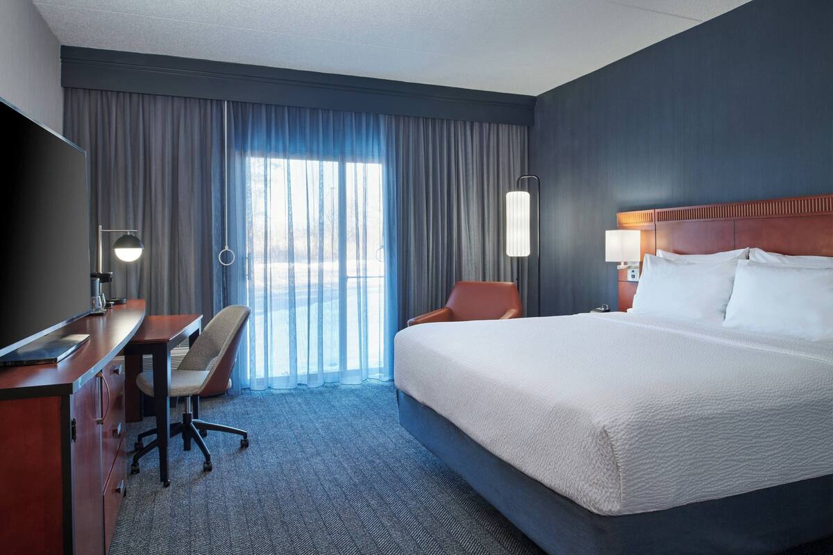 A hotel room with a large bed, a desk with a chair, a TV, and a window with curtains.