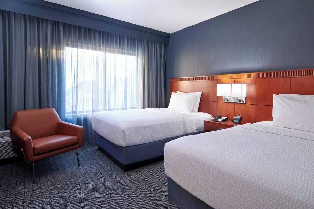 A hotel room with two beds, a brown leather chair, a nightstand with a phone, and a large window with curtains.