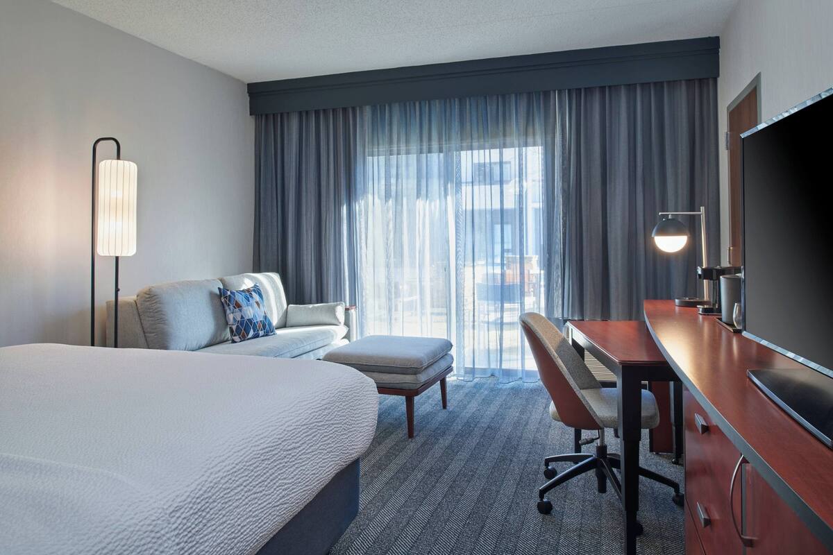 A hotel room with a large bed, a desk with a chair, a sofa, a standing lamp, and a large TV.
