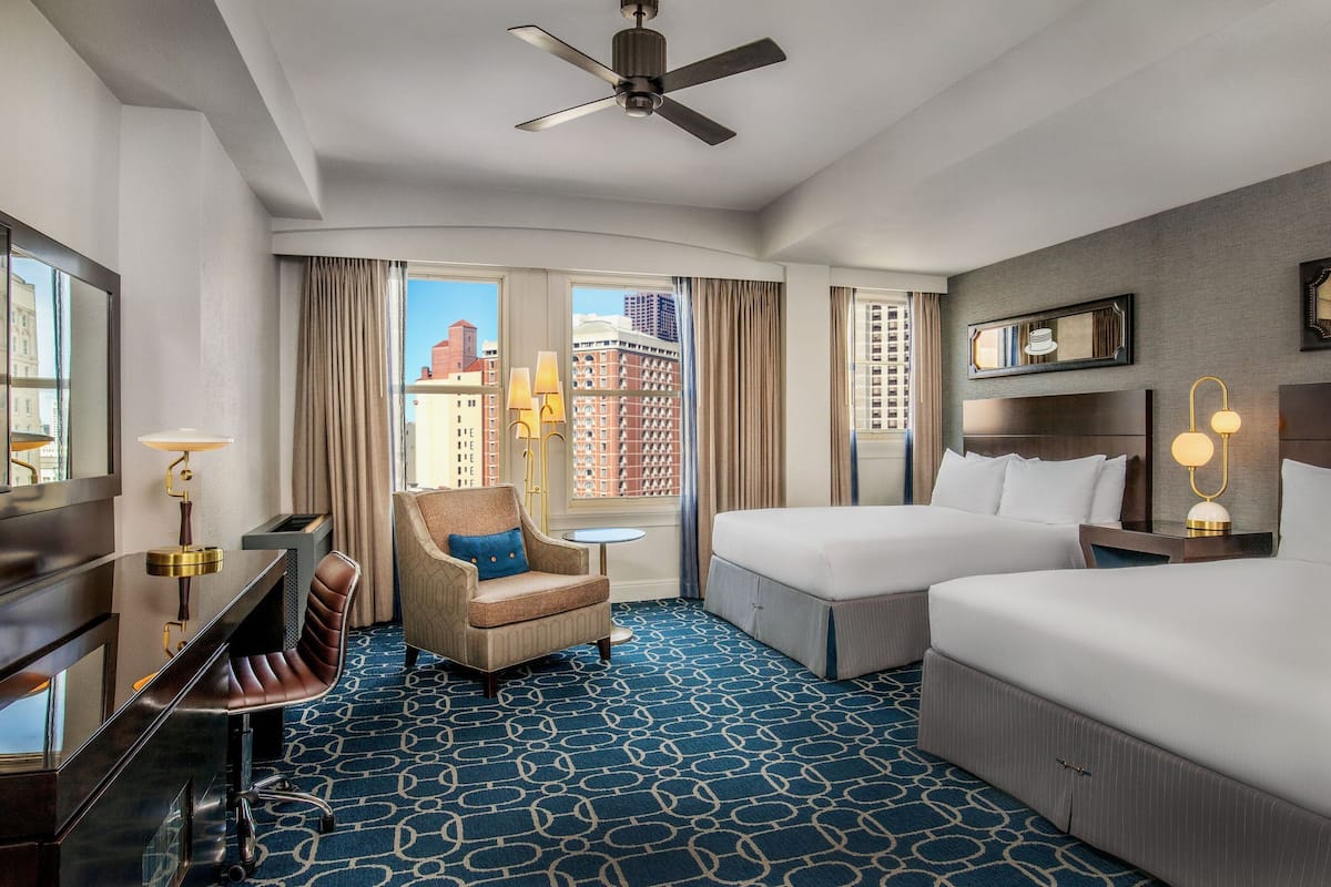 A hotel room with a bed, a desk, a chair, a small table, and a view of buildings through the window.
