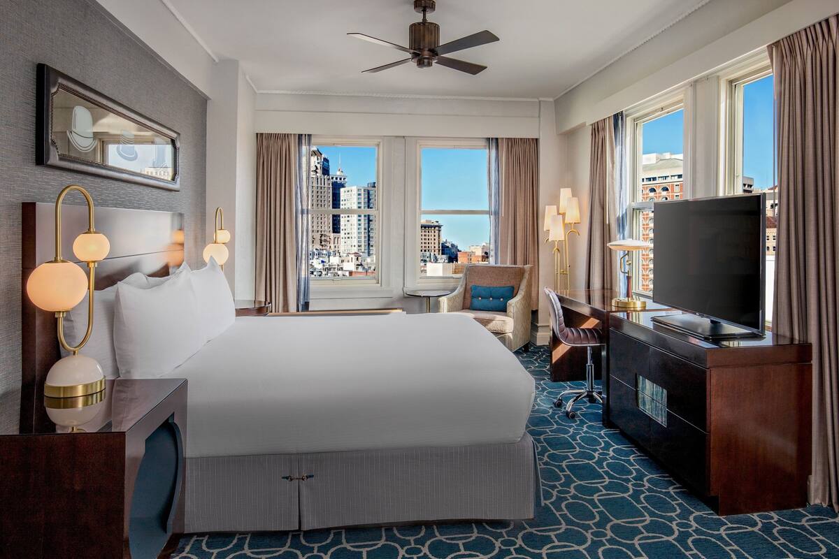 A hotel room with a large bed, a desk with a TV, a chair, a small table, and a view of the city through large windows.