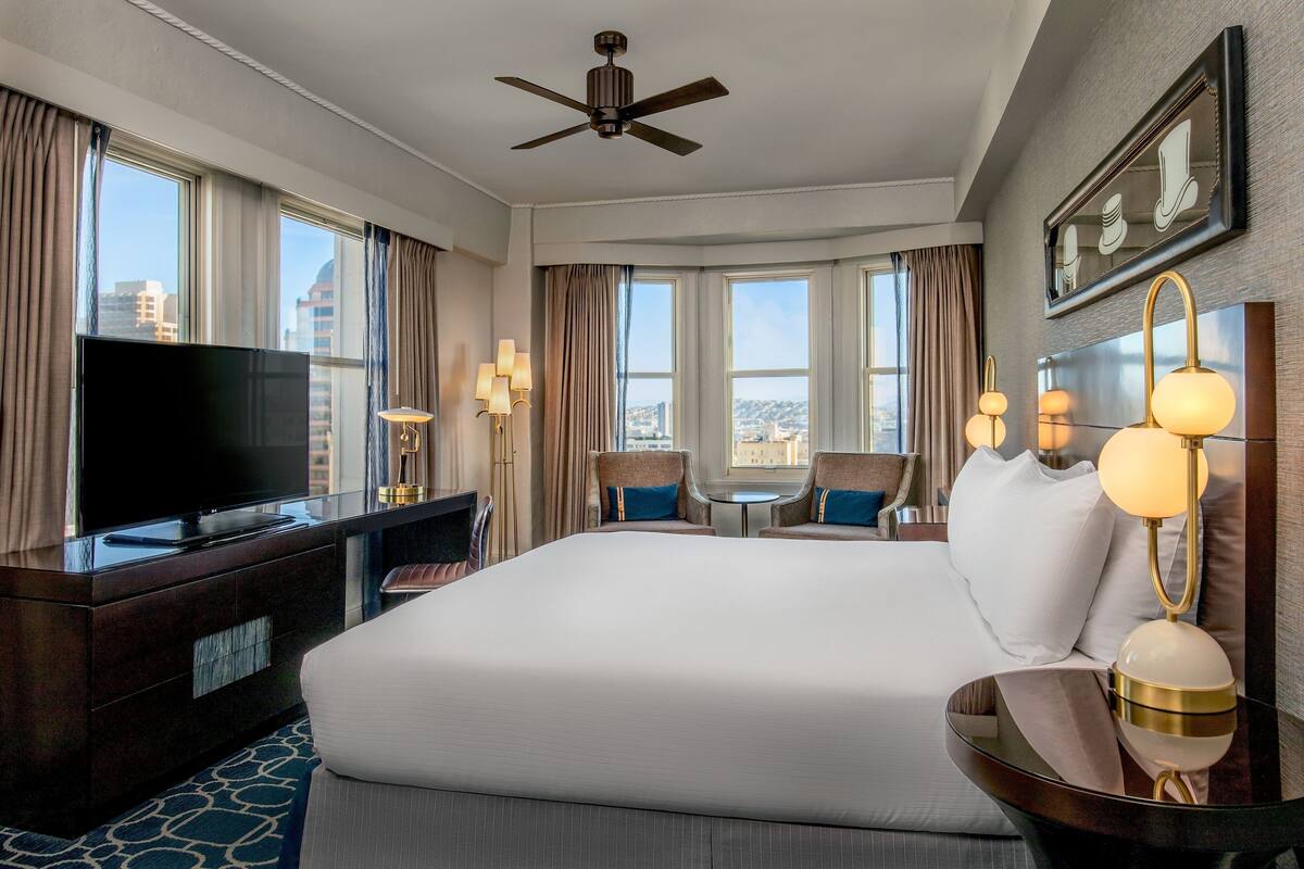 A hotel room with a large bed, a flat-screen TV, a desk with a lamp, and a view of the city through large windows.