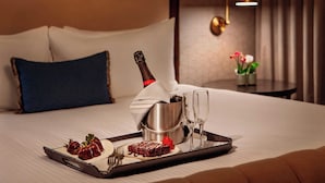 Un lit bien fait, avec un plateau de desserts et une bouteille de champagne, une table de chevet avec une lampe et un vase rempli de fleurs.
