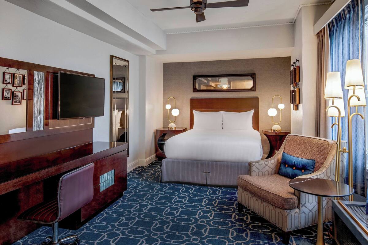 A hotel room with a large bed, a desk with a flat-screen TV, a chair, and a small table.