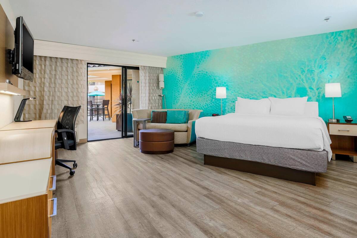 A modern hotel room with a large bed, a desk with a chair, a small round ottoman, and a television mounted on the wall.