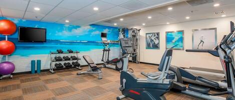 Ein Fitnessstudio mit Trainingsgeräten, einem an der Wand befestigten Fernseher und einem Wandbild mit einer Stranddarstellung.