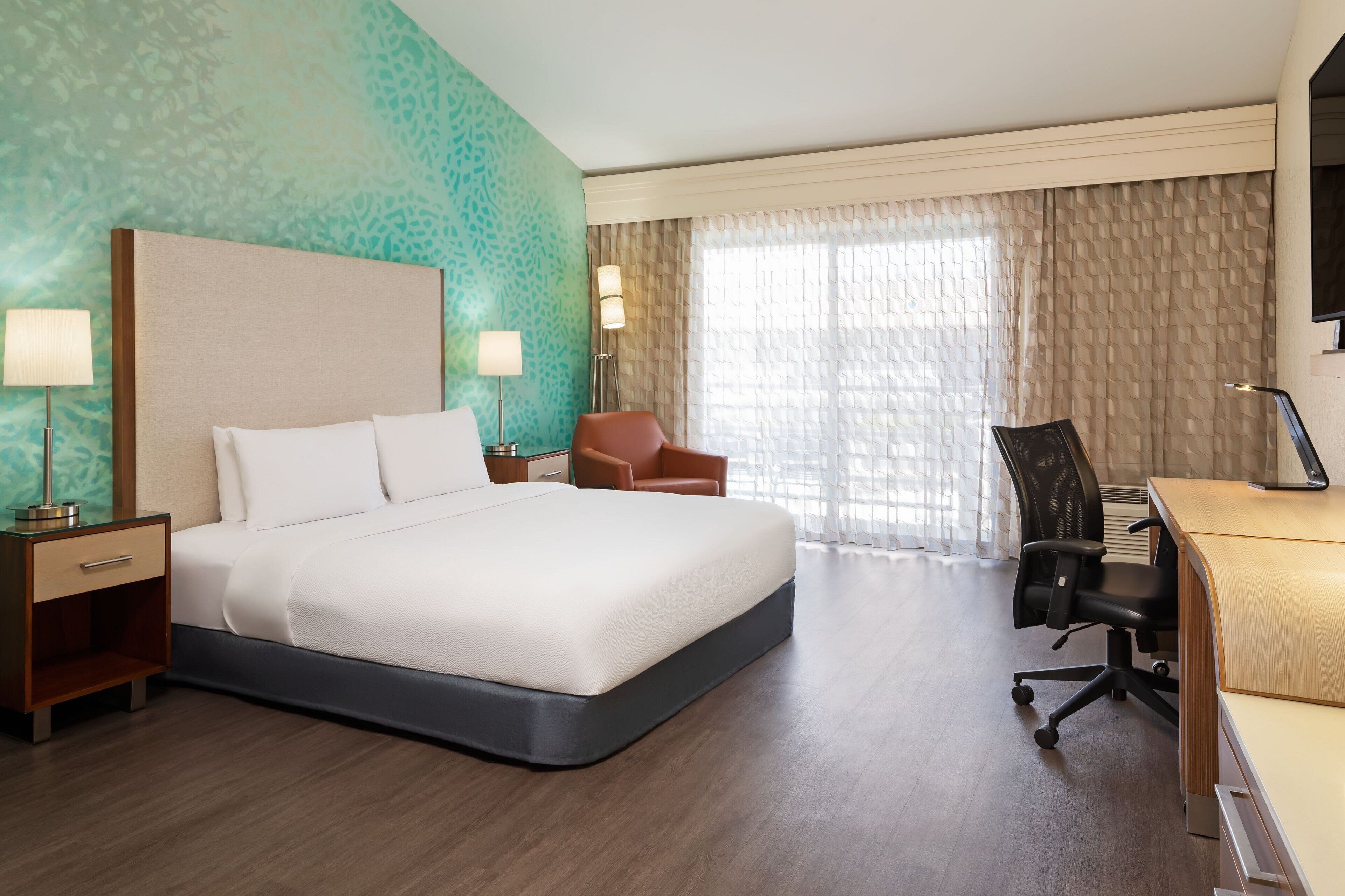 A hotel room with a large bed, a desk with a computer, a chair, and a lamp.