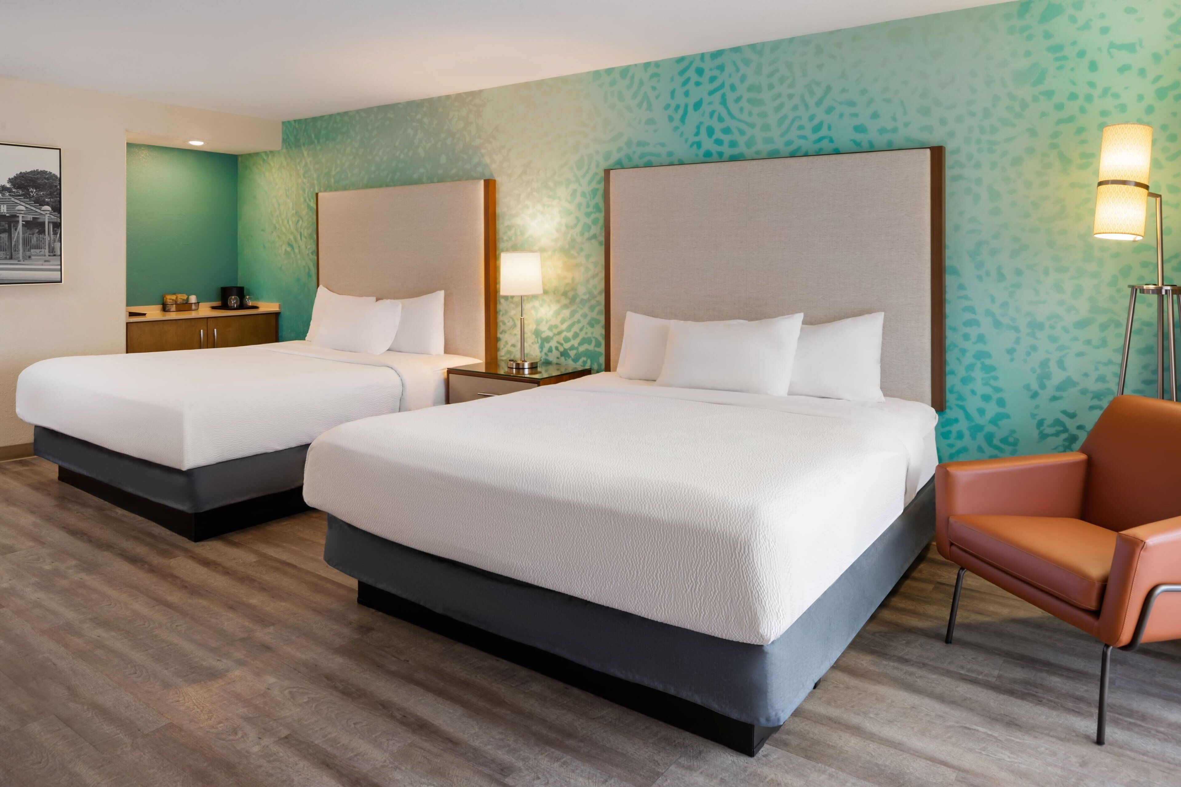 A hotel room with two beds, a nightstand, a chair, and a wall-mounted lamp.