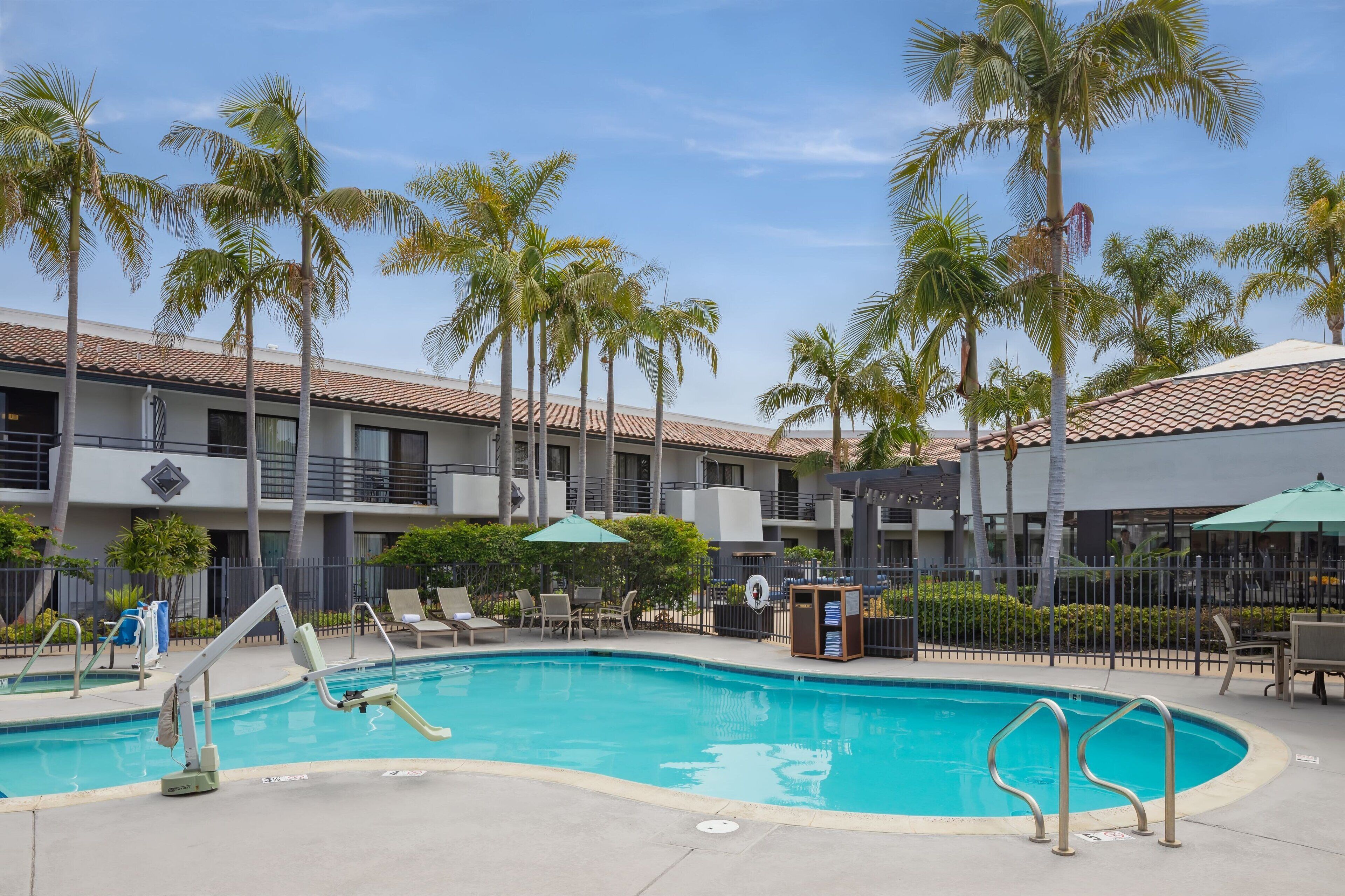 A swimming pool with lounge chairs and umbrellas, surrounded by palm trees and a building with a tiled roof.
