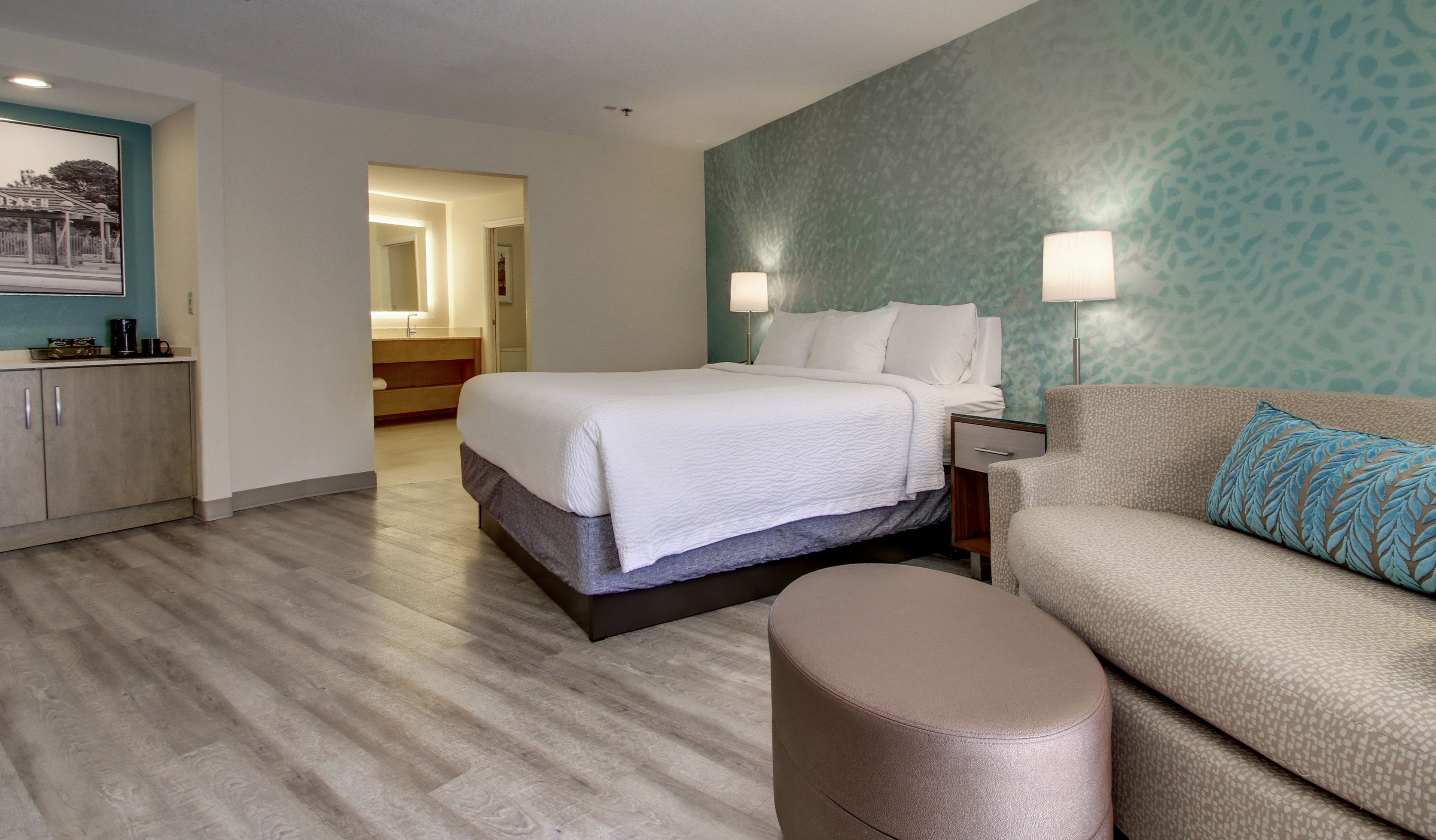 Une chambre d’hôtel comprenant un lit, un canapé, un petit pouf, une table de chevet et une salle de bain visible à travers une porte ouverte.
