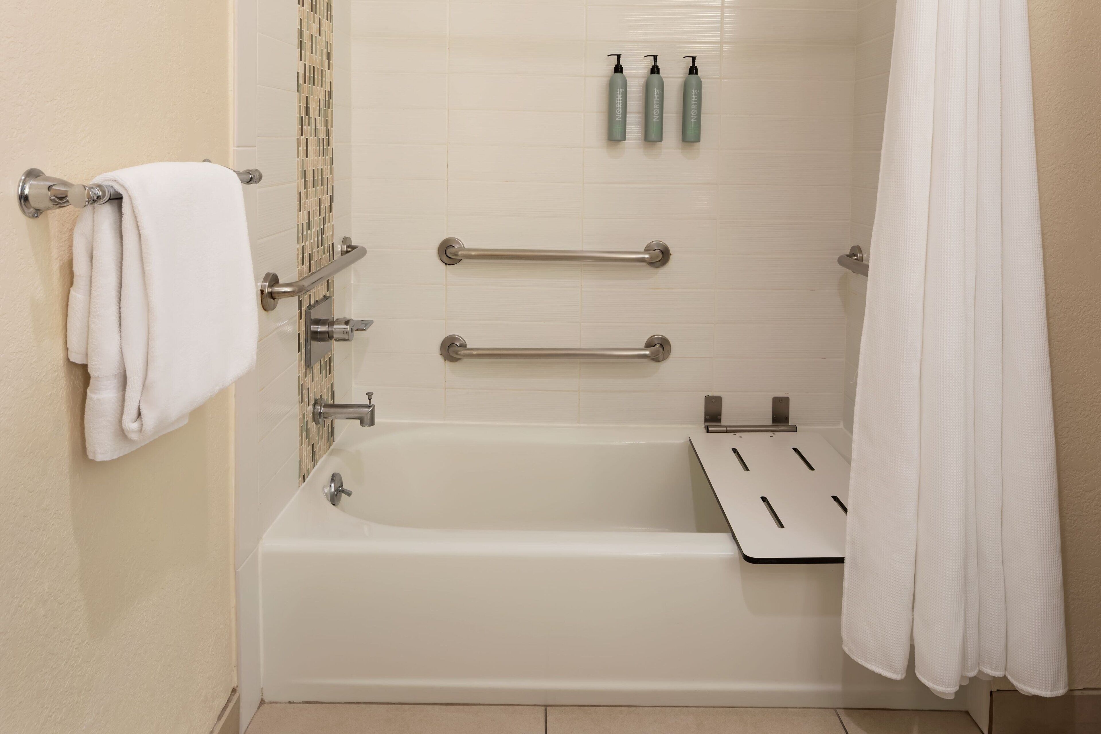 A modern bathroom with a bathtub, grab bars, a towel rack, and toiletries on the wall.
