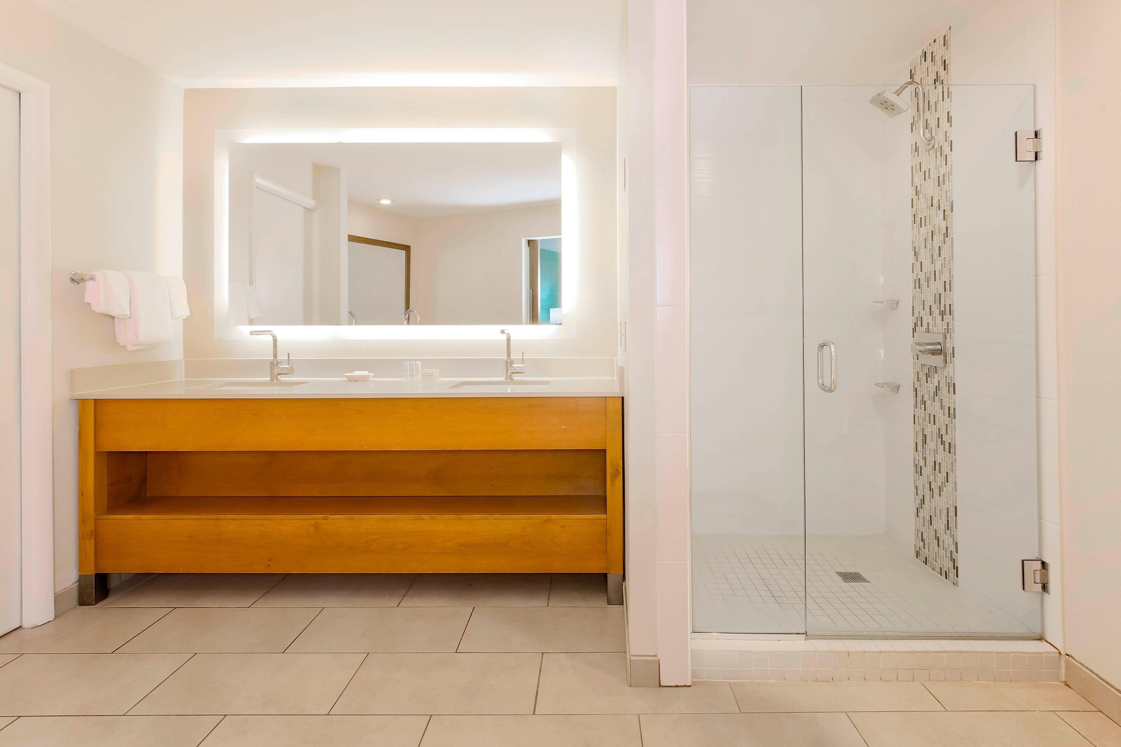 A modern bathroom with a large mirror, a wooden vanity, a glass shower enclosure, and a tiled floor.