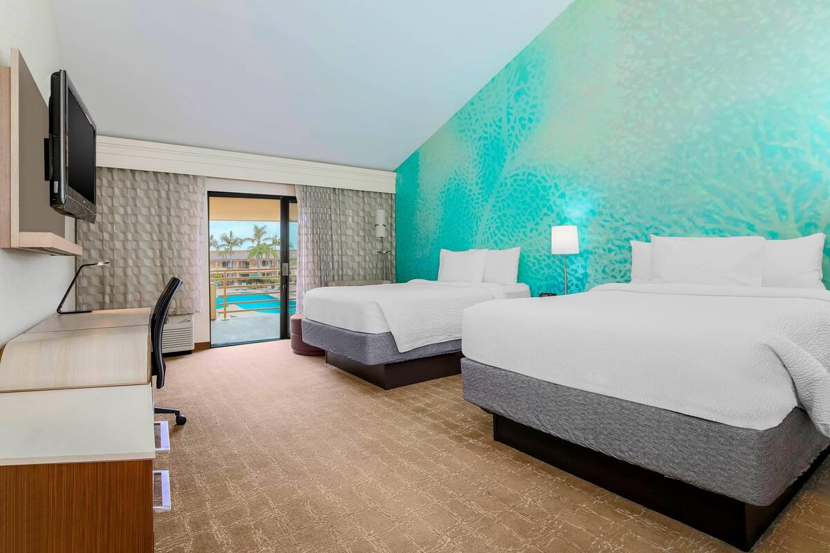A hotel room with two beds, a desk, a TV, and a view of a pool.