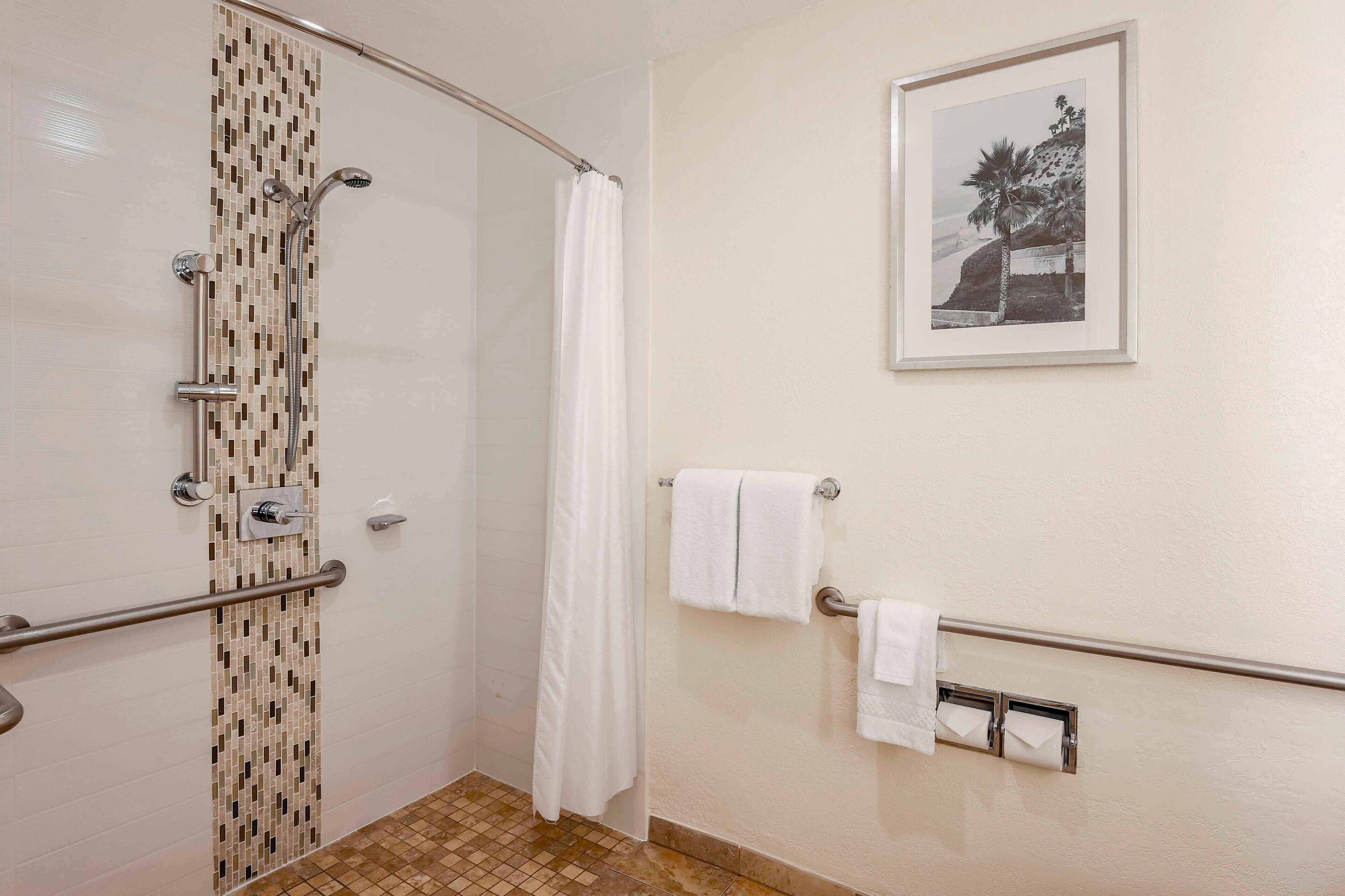 A shower with a tiled wall, a glass door, a towel rack with two towels, and a framed picture on the wall.