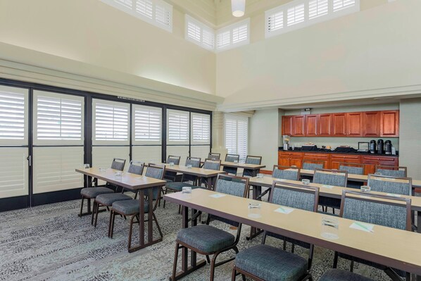Un salón de conferencias con varias mesas y sillas, ventanas amplias con persianas y una zona de cocina con armarios de madera.
