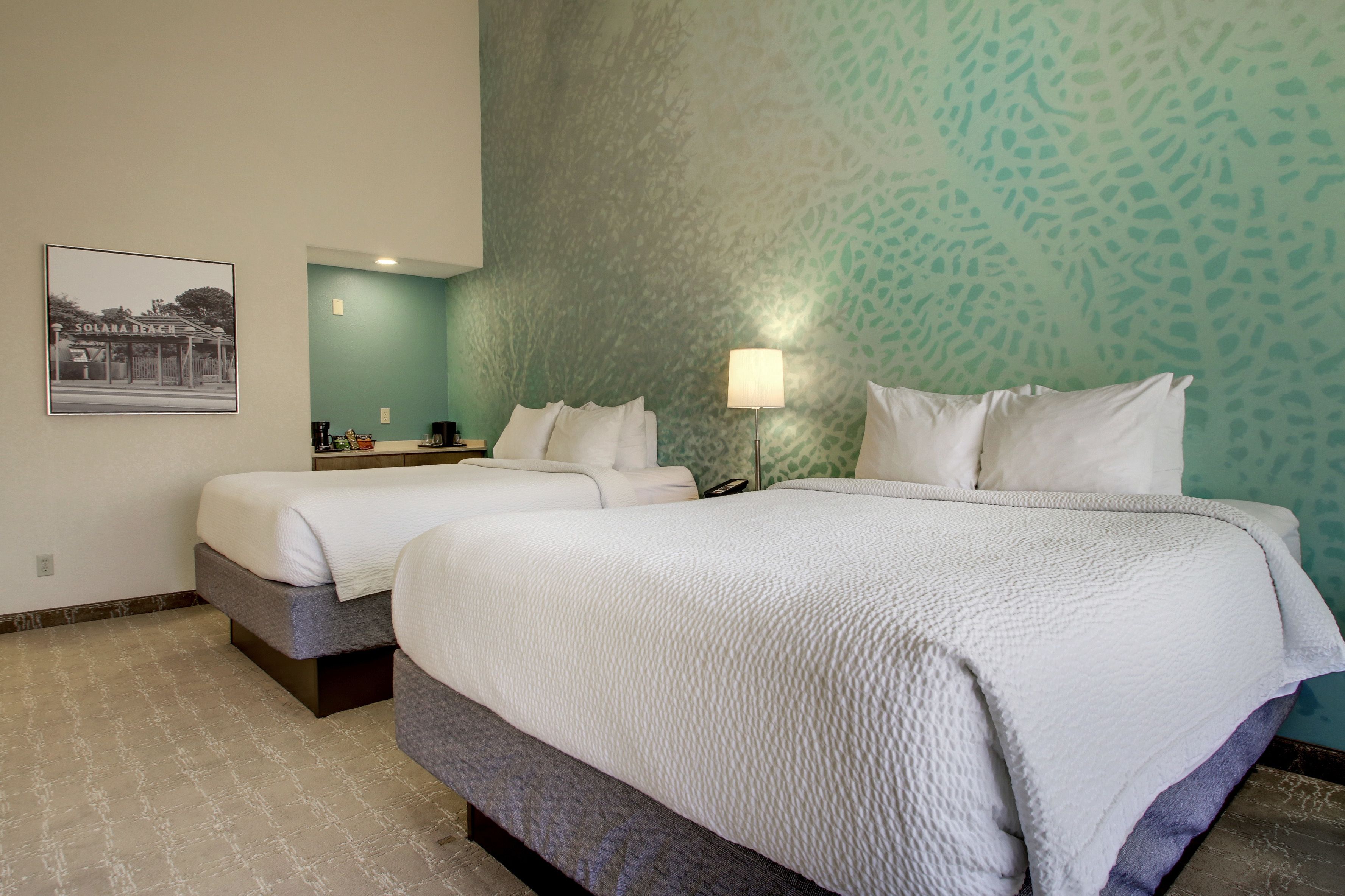 A hotel room with two beds, a teal patterned wall, a framed picture on the wall, and a small table with a lamp.