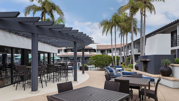 An outdoor patio area with tables and chairs, a pool, and palm trees. - Courtyard by Marriott San Diego Del Mar/Solana Beach (Solana Beach)